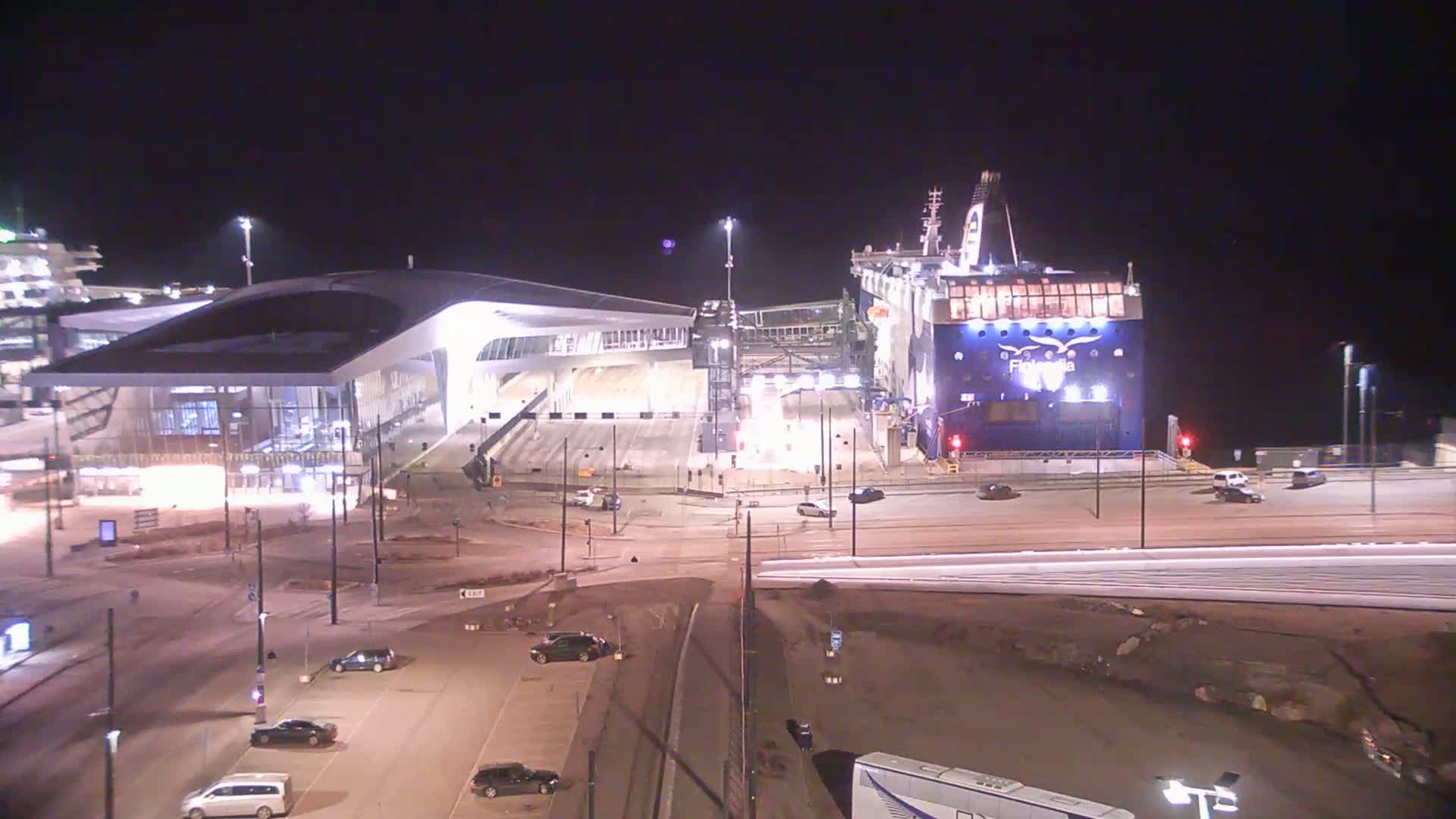 At night, a large ferry is docked at a terminal building, with several vehicles visible in the surrounding parking areas and roadways.