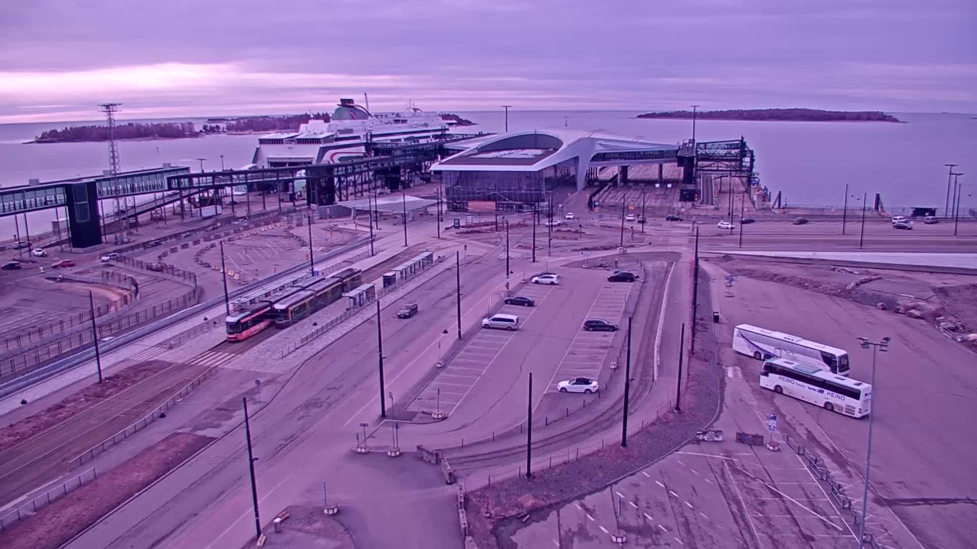 An aerial view shows a ferry terminal with a ferry docked, a tram, several cars in a parking lot, and two buses, all under a dusky, overcast sky.