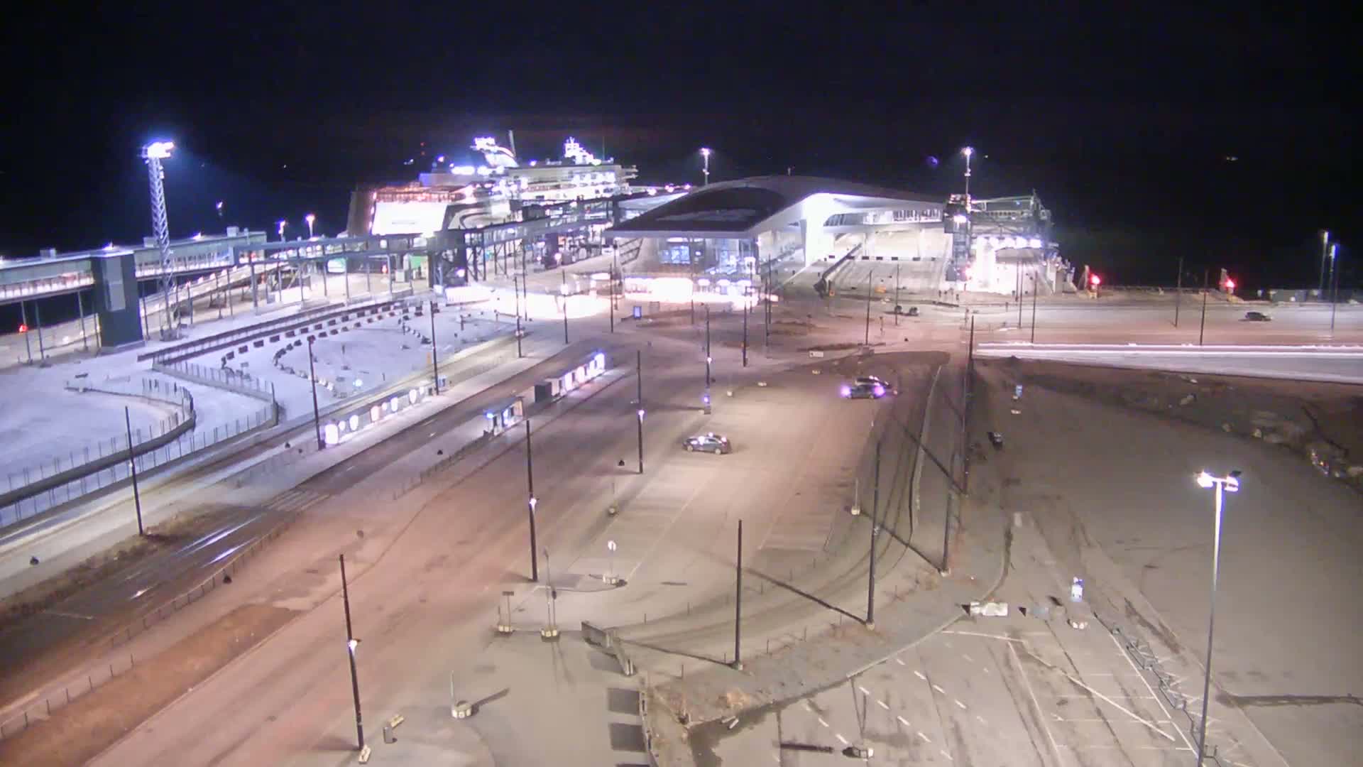 A nighttime aerial view of a ferry terminal with a large ferry docked, surrounded by roadways, parking areas, and some light snow on the ground.