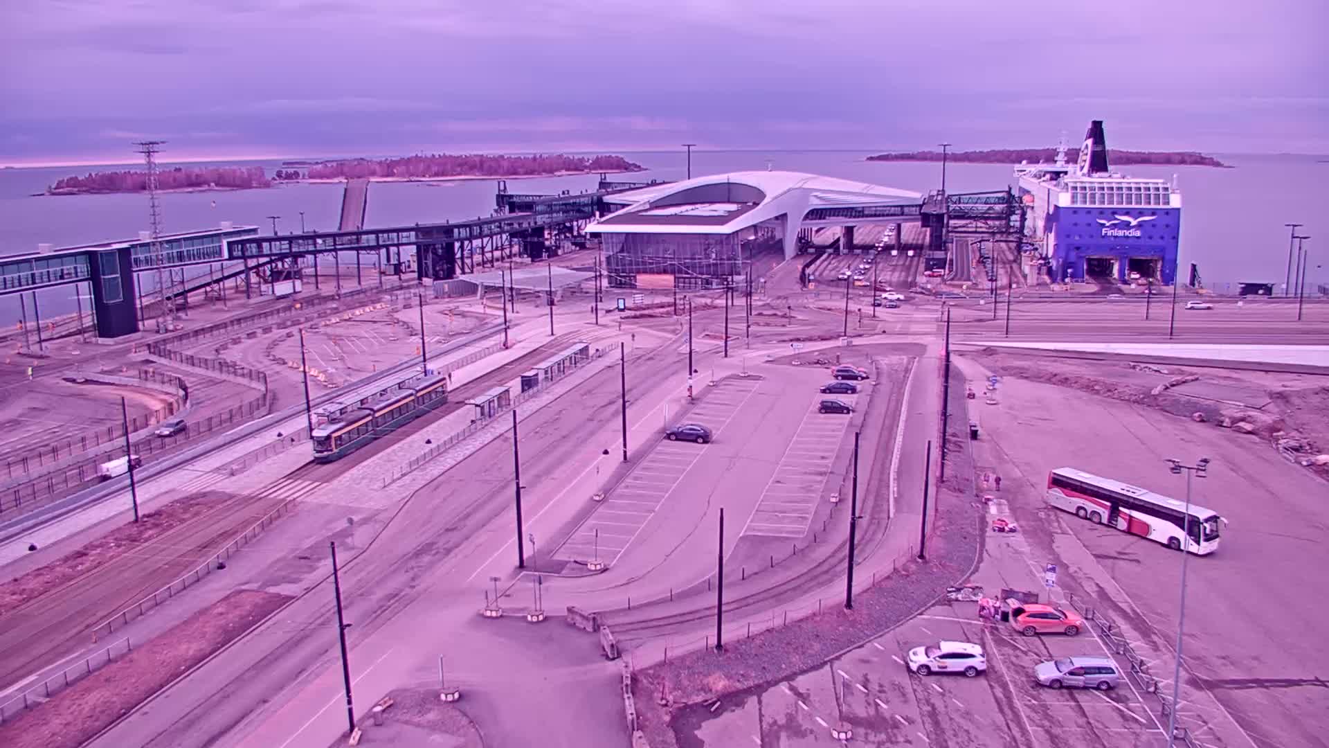 An aerial view of a ferry terminal under a pale purple sky shows a large ferry docked, a tram on tracks, several cars in a parking lot, and a bus parked nearby.