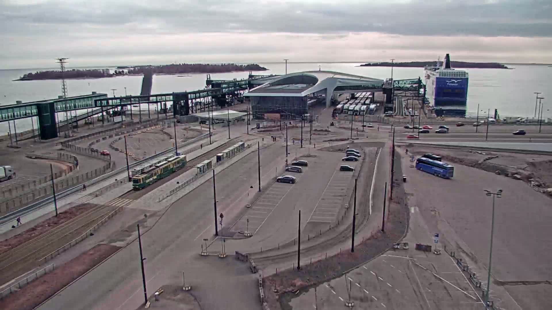 An aerial view shows a ferry terminal on an overcast day, with a ferry docked, vehicles in parking lots, a tram on the tracks, and a bus on the road.