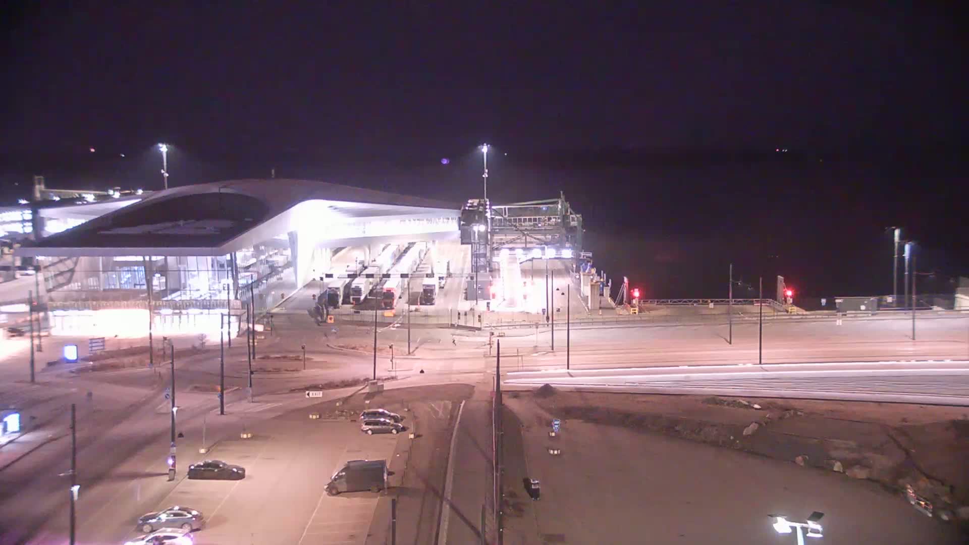 A brightly lit ferry terminal at night with a few cars parked nearby, under a dark, clear sky.