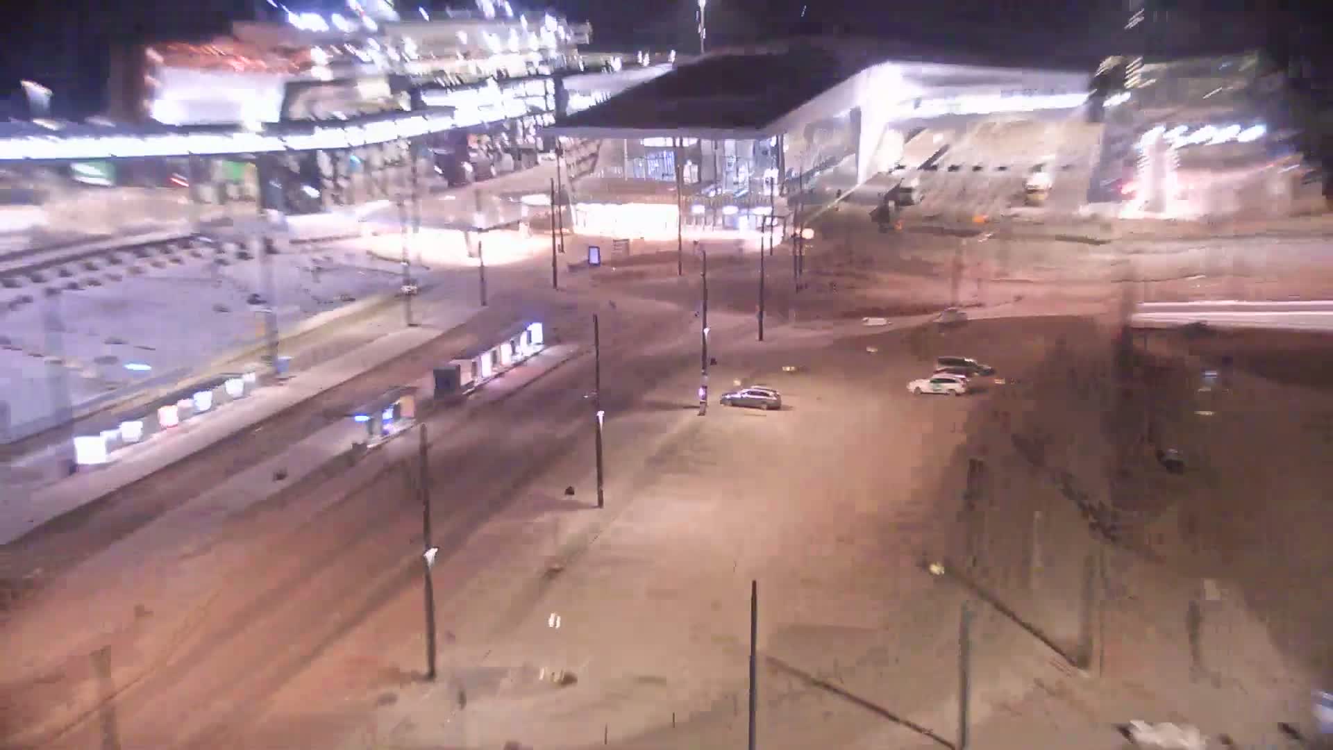 A nighttime aerial view of a snow-covered city square with a train station, tram tracks, and a few cars parked.