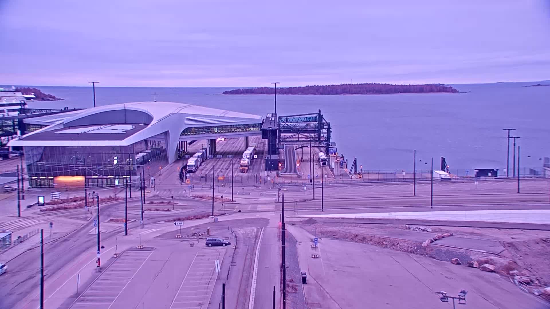 A ferry terminal with several vehicles, a modern building, and a calm body of water under a pale purple sky.