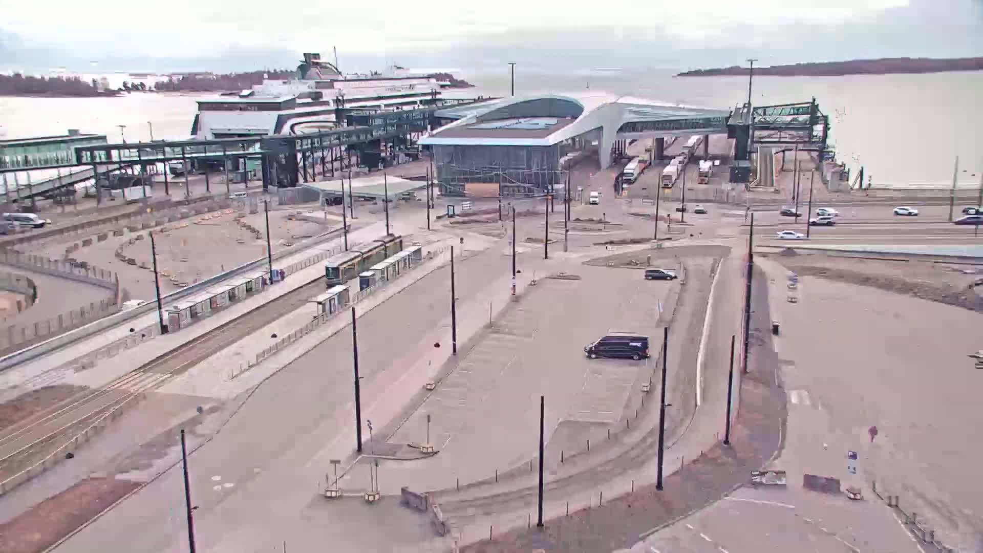 An aerial view shows a ferry terminal with a tram station, a large parking lot, and several vehicles on a cloudy day.