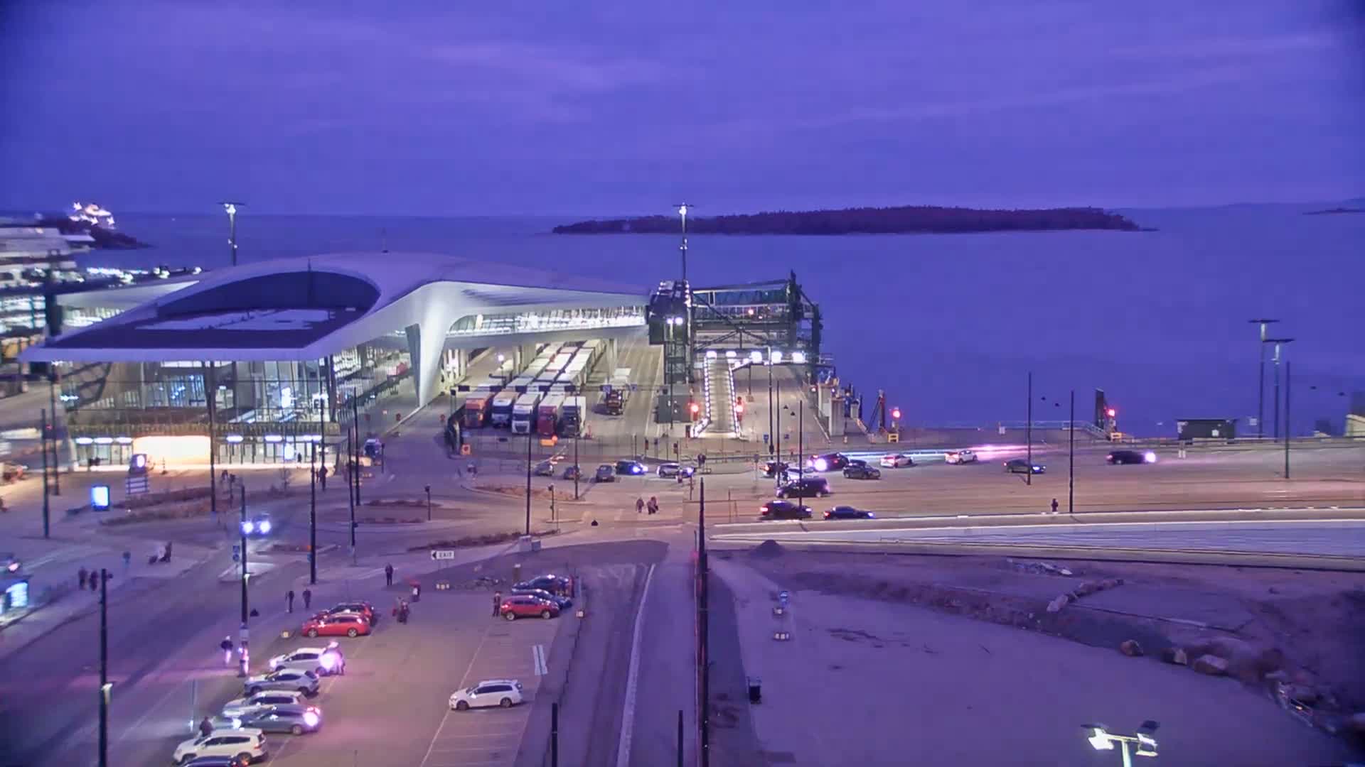 A modern ferry terminal with numerous vehicles and buses is situated by a calm body of water under a twilight sky.