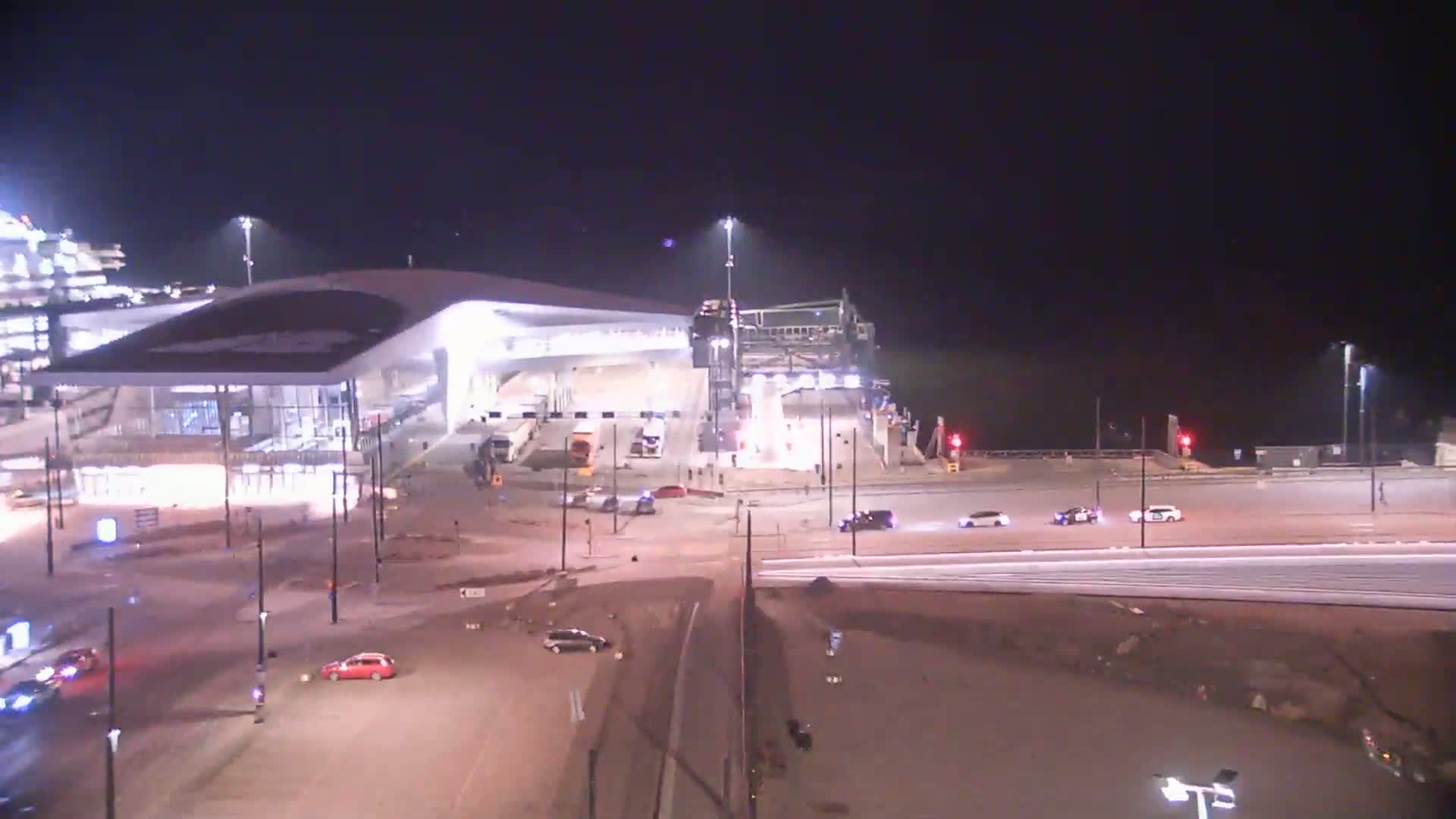 At night, a brightly lit modern building with a curved roof is partially under construction, with vehicles moving around it on a mostly empty road.
