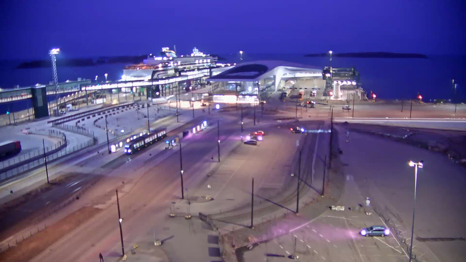 A ferry terminal at dusk, with a large ferry docked, a tram on the road, and several cars parked in an adjacent lot under a twilight sky.