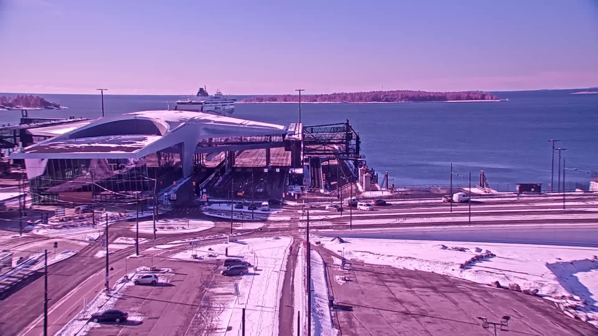 A ferry terminal with a modern building and a partially snow-covered parking lot overlooks a calm body of water on a sunny day.