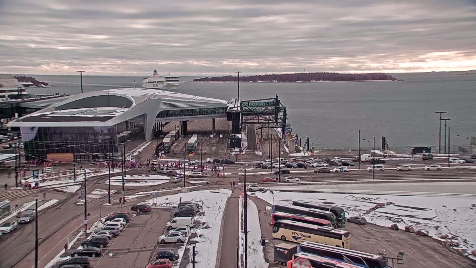 A ferry terminal with a modern building, numerous cars and buses in a snowy parking lot, and a ferry visible in the background on a cloudy day.