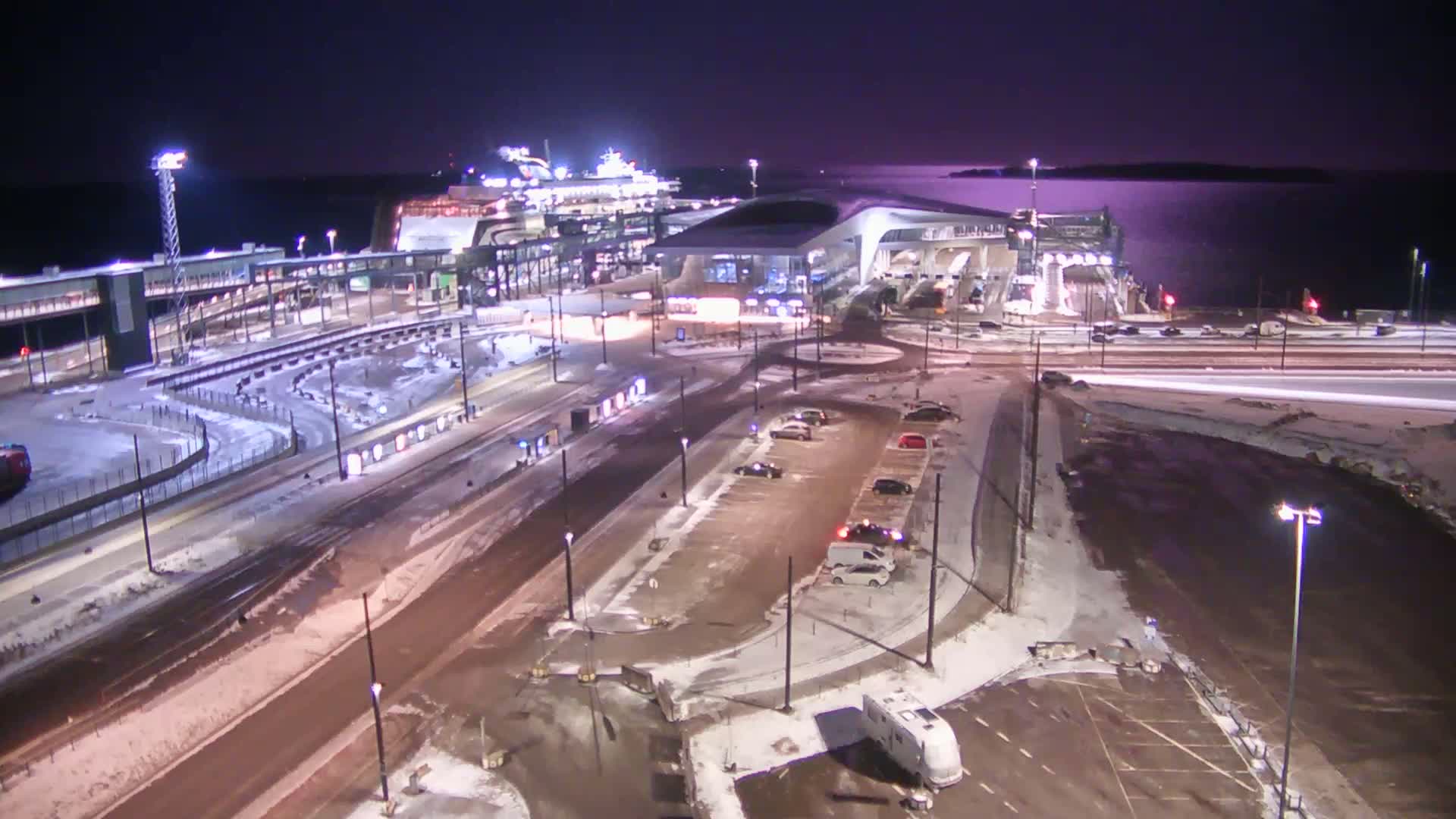 A cruise ship is docked at a terminal at night in snowy conditions, with cars parked in nearby lots and roads partially covered in snow.