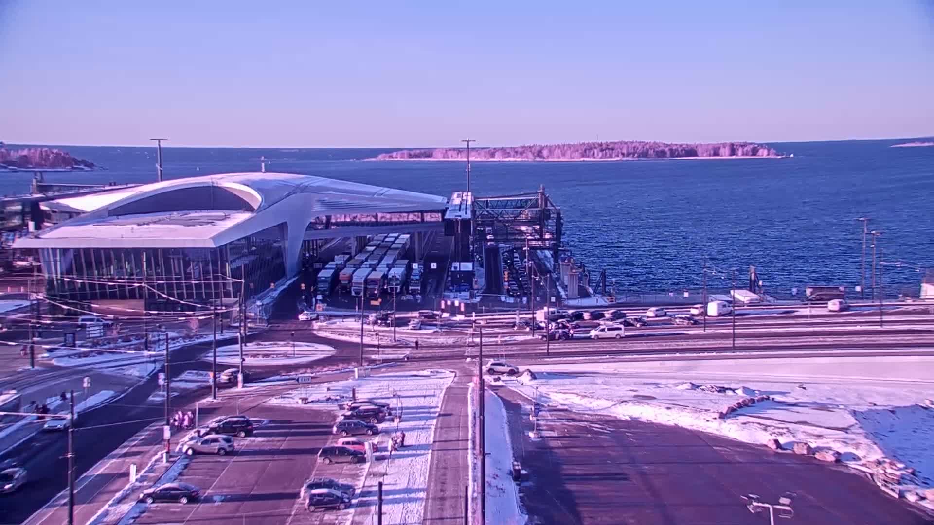 A ferry terminal with several vehicles and trucks, partially covered in snow, sits on a waterfront with calm blue water and a small island in the distance under a clear, bright sky.