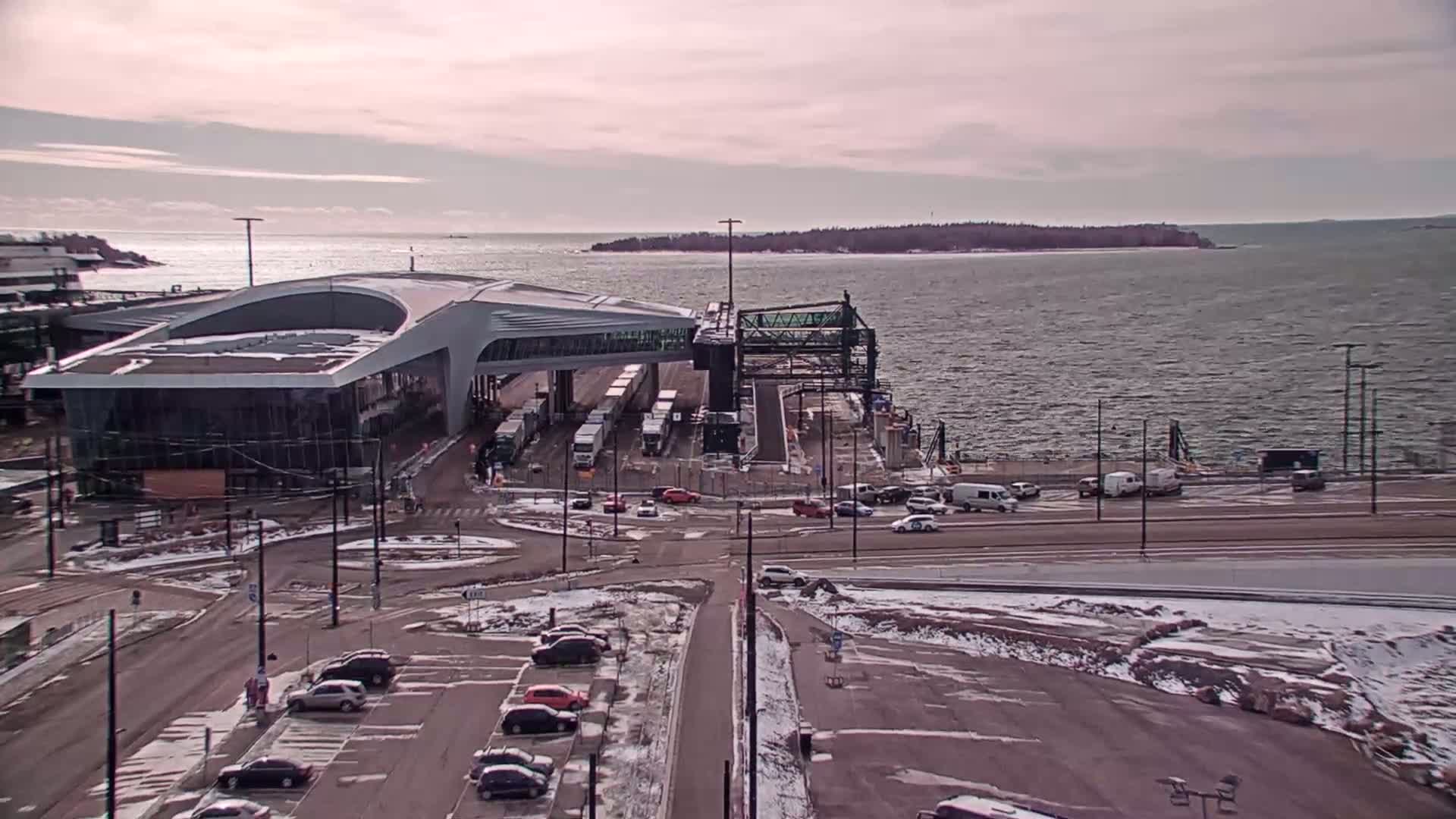 A ferry terminal with a modern building and several vehicles is seen on a partly cloudy day, next to a body of water with an island in the distance.