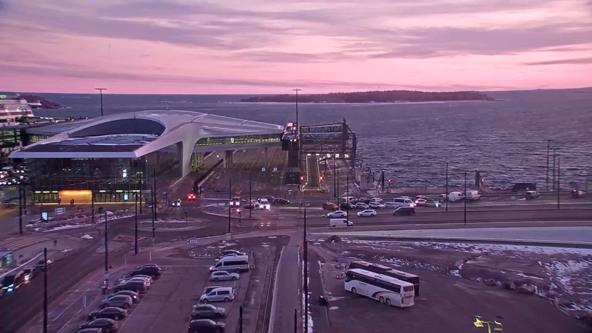A modern ferry terminal with cars and a bus parked nearby overlooks a wavy sea under a pinkish sunset.