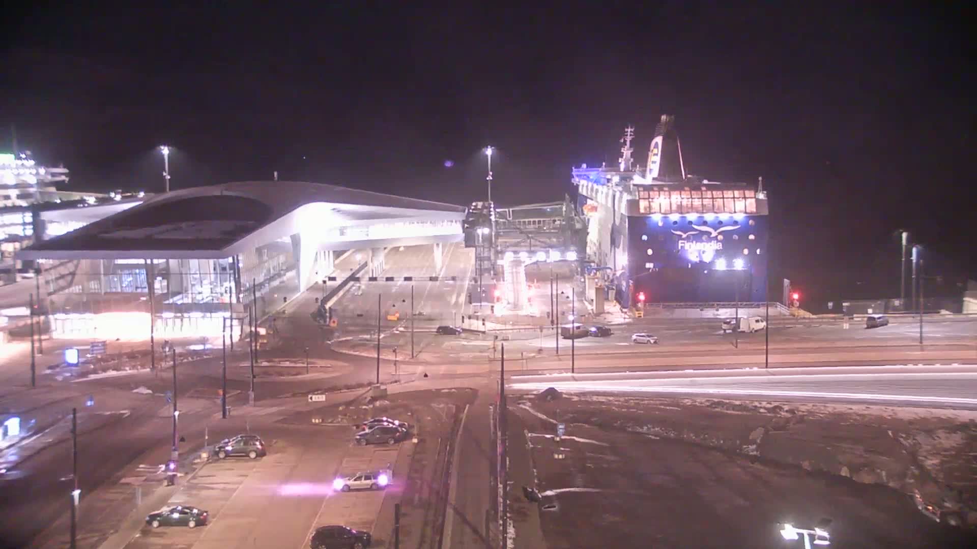 A large ferry is docked at a terminal at night, with cars parked nearby and light snow or slush on the ground.