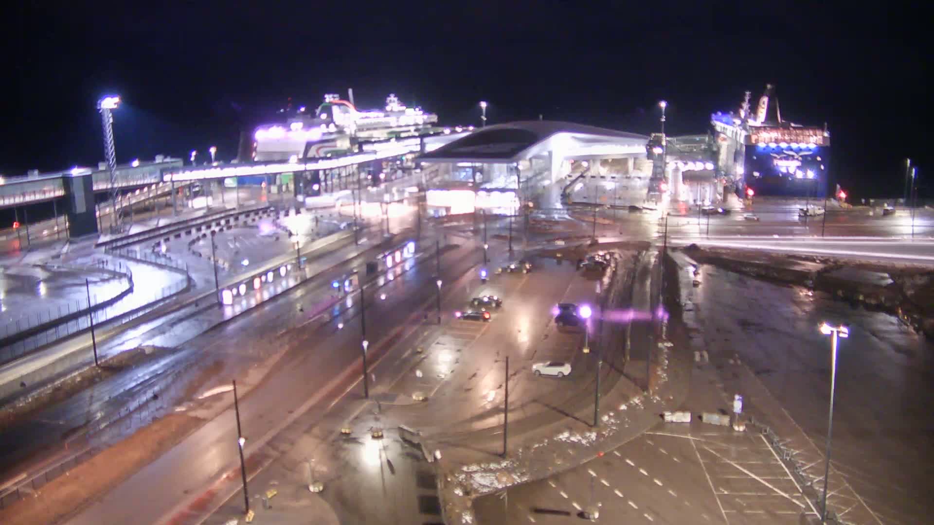 A nighttime, rain-soaked ferry terminal with two large ferries docked, surrounded by wet roads and parking areas with a few vehicles.