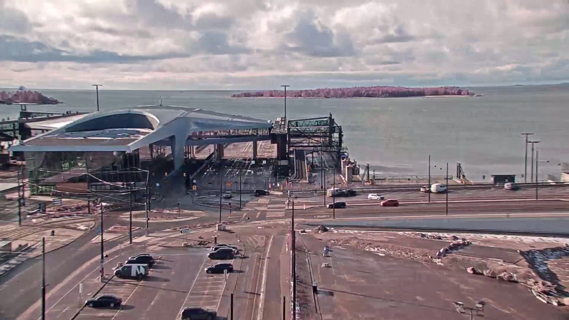 A ferry terminal with a modern building and parking lot overlooks a body of water under a partly cloudy sky.