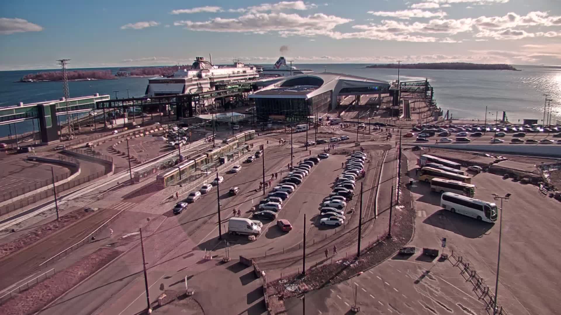 A ferry terminal on a sunny day with a partly cloudy sky, showing numerous cars and buses parked, a tram, and a large ferry docked.