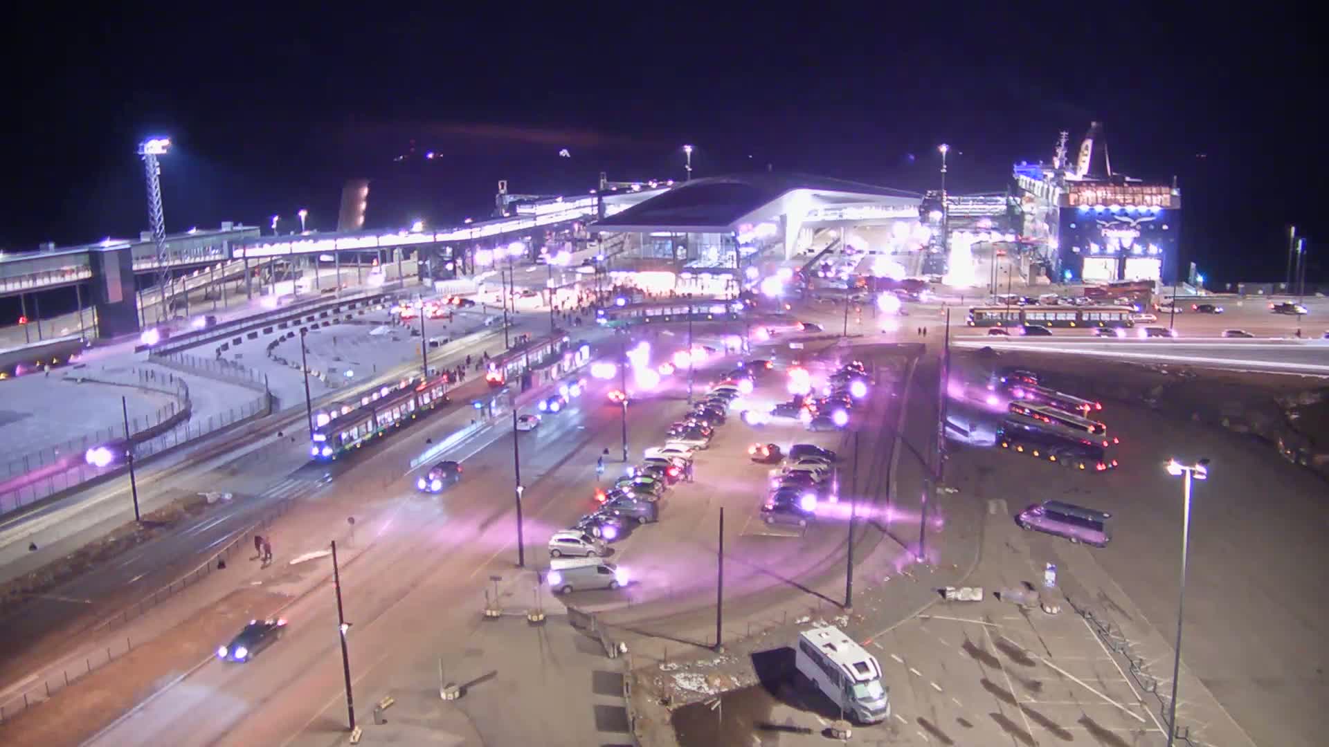 A nighttime aerial view shows a large terminal with numerous vehicles, including buses and cars, parked in lots and on roadways; a tram is also visible, with purple light effects emanating from various locations.