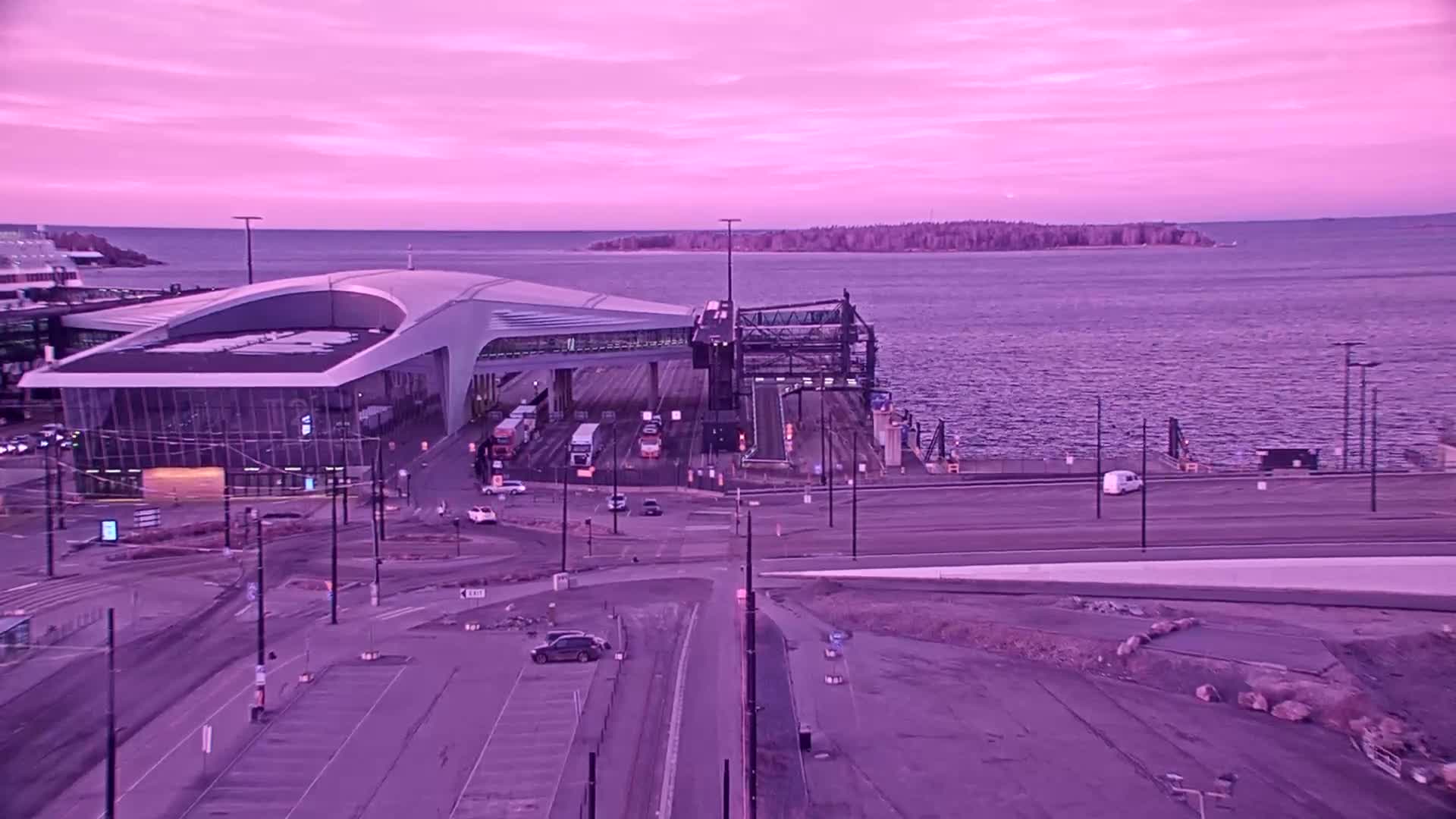 A ferry terminal with several vehicles is situated on a waterfront under a purplish-pink sky at dawn.