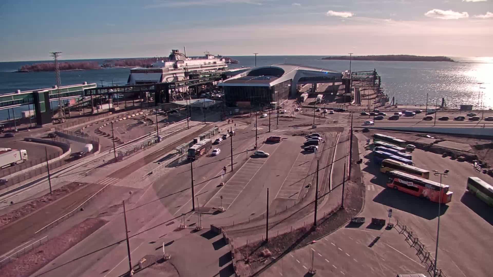 An aerial view shows a ferry terminal on a sunny day, with several buses and cars parked nearby, and a large ferry docked at the terminal.