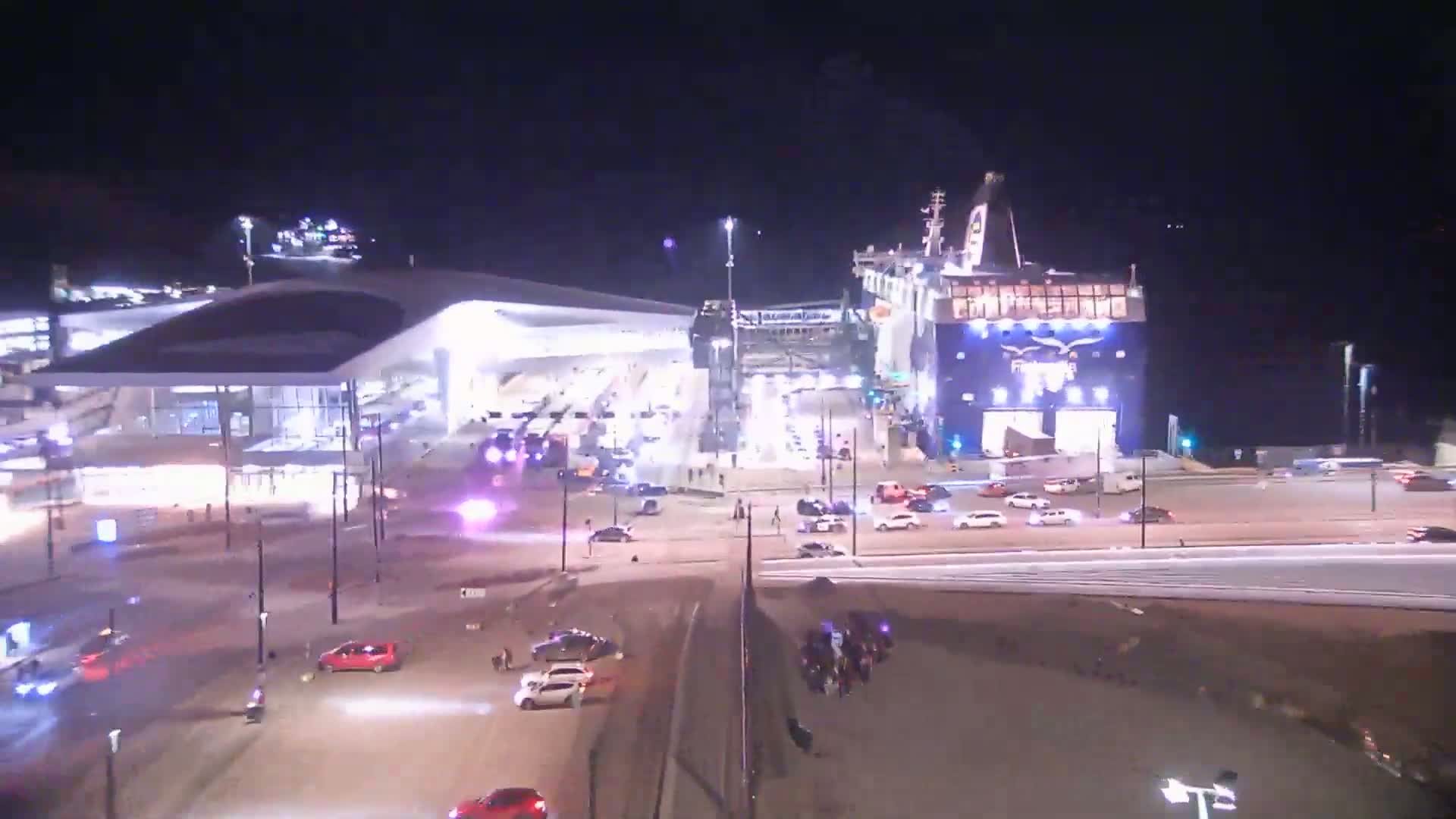 At night, a large ferry is docked at a terminal with cars driving around the area.