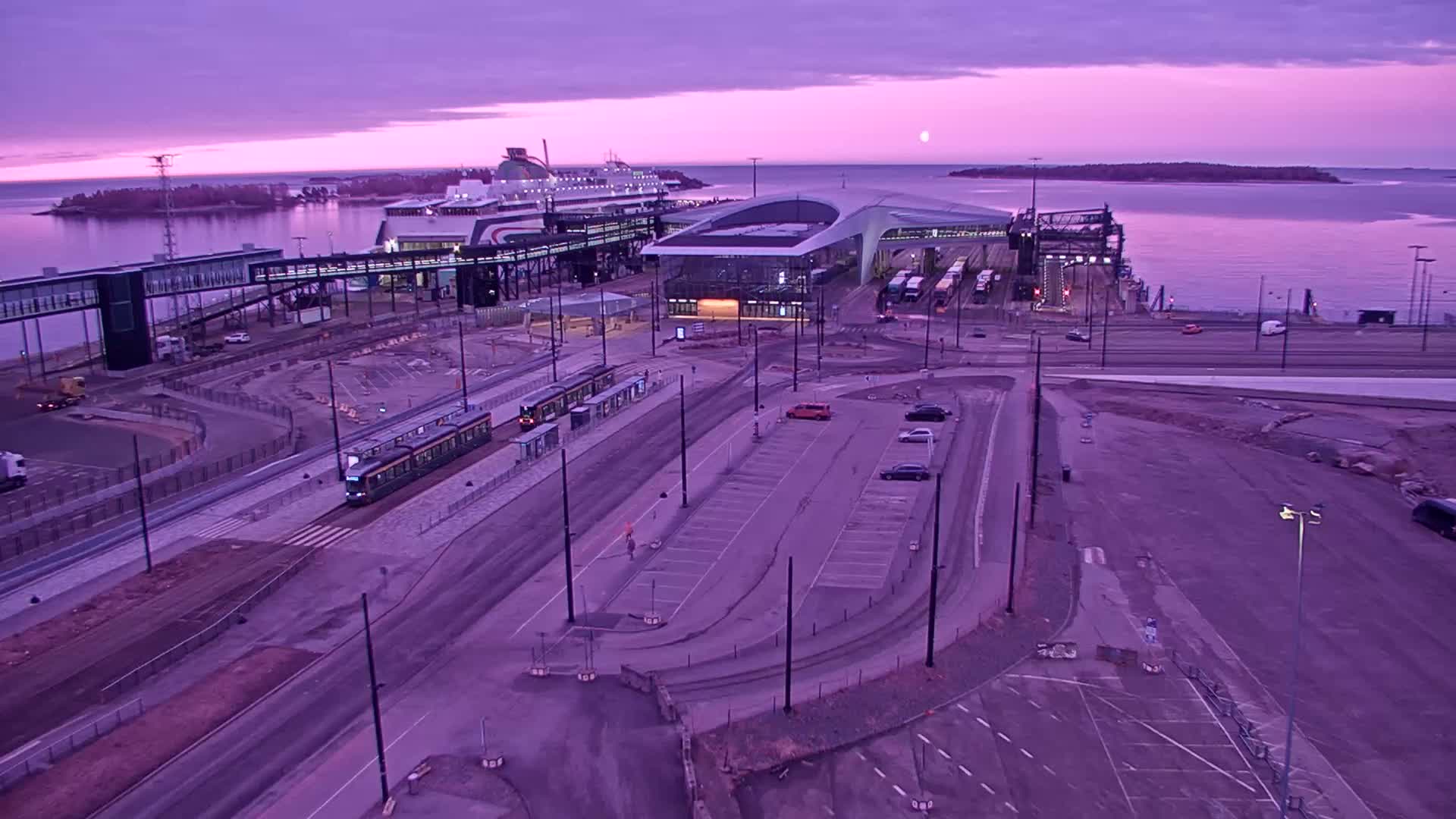 An aerial view of a ferry terminal at sunrise under a purple sky, showing a tram, cars in a parking lot, and a large ferry docked.