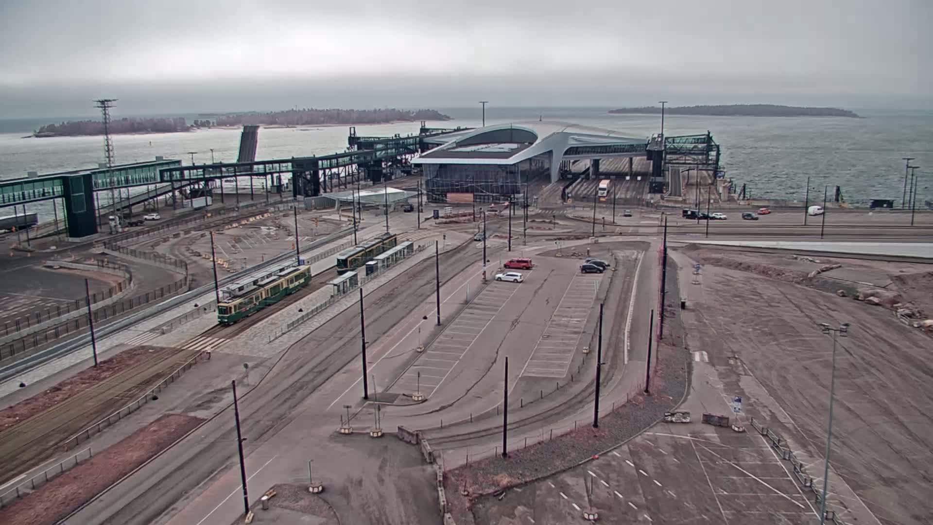 An aerial view shows a ferry terminal with trams and cars parked near a large building, under an overcast sky.