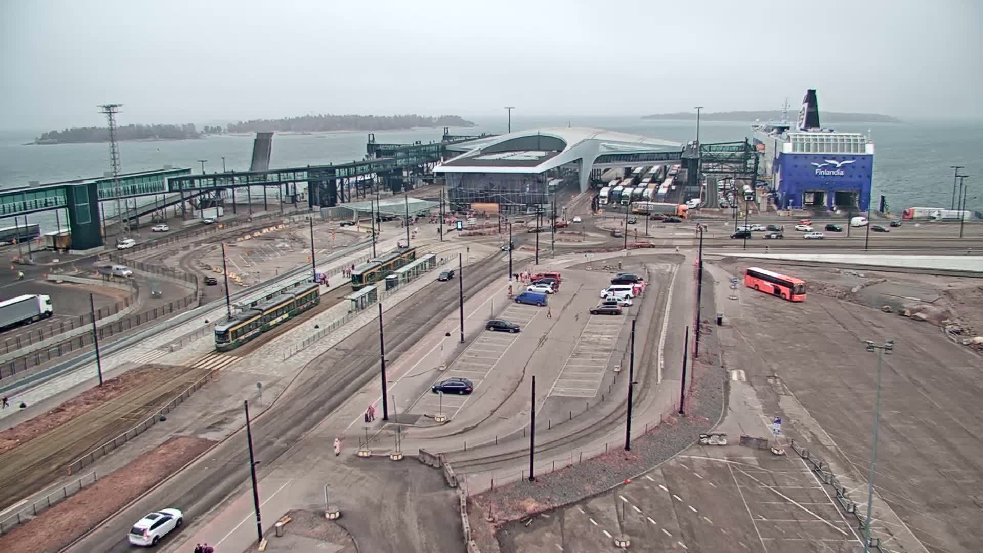 A ferry is docked at a terminal with trams and vehicles in a parking lot under an overcast sky.