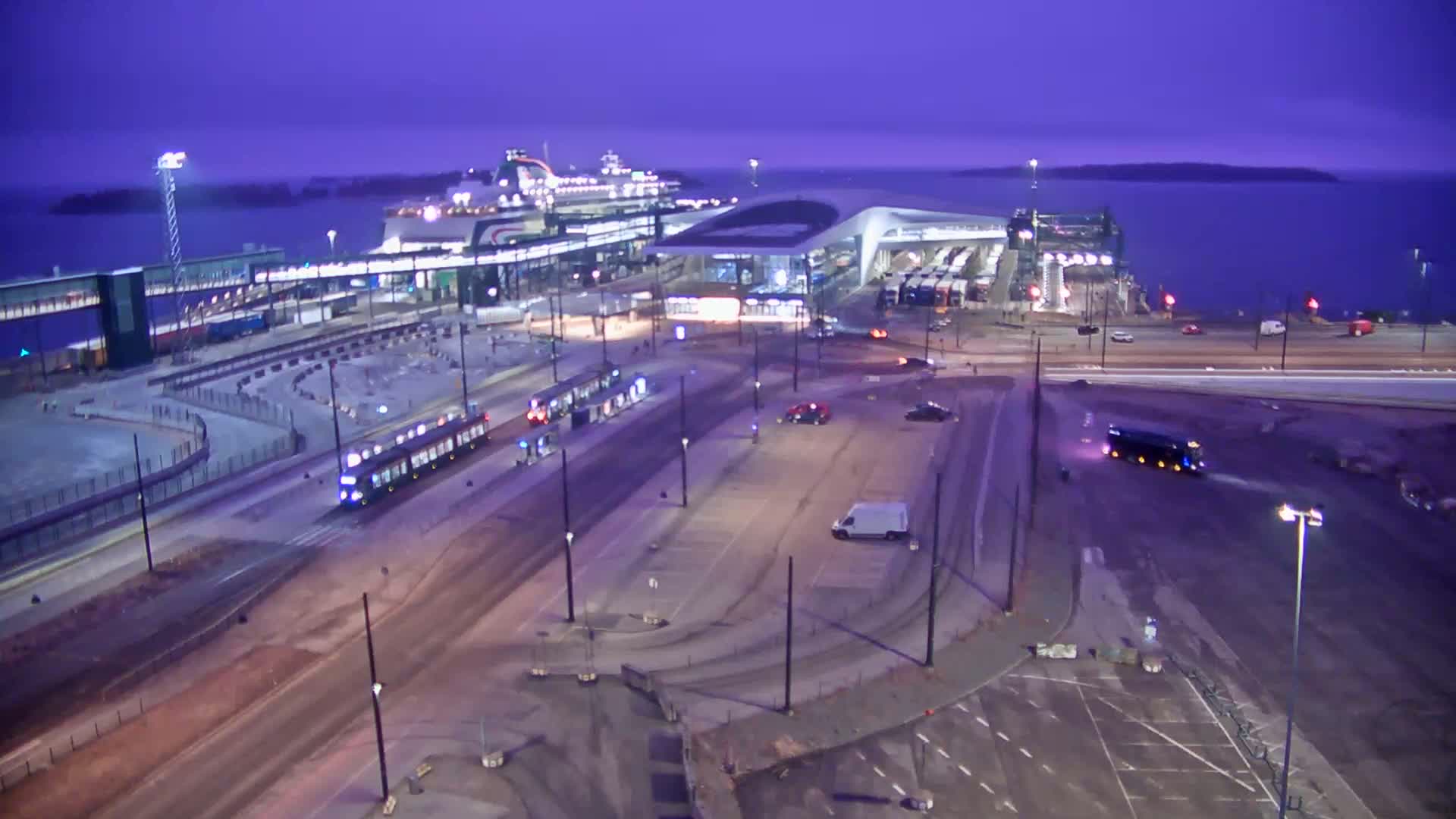An aerial view at dusk shows a ferry docked at a terminal, with trams and vehicles in a surrounding area undergoing some construction.