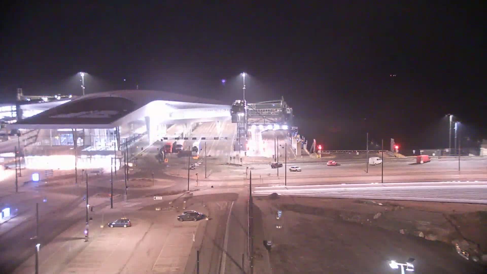 A brightly lit ferry terminal at night, with several vehicles parked nearby, under a dark sky.