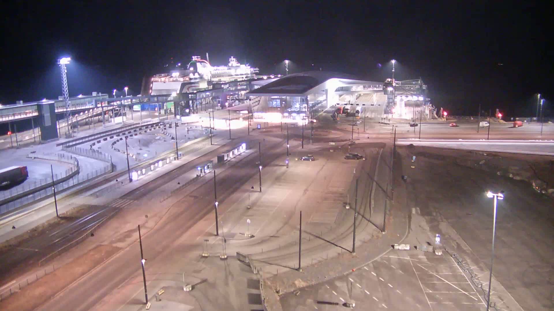 A large ferry is docked at a brightly lit terminal at night, with an empty, partially paved area surrounding the terminal.