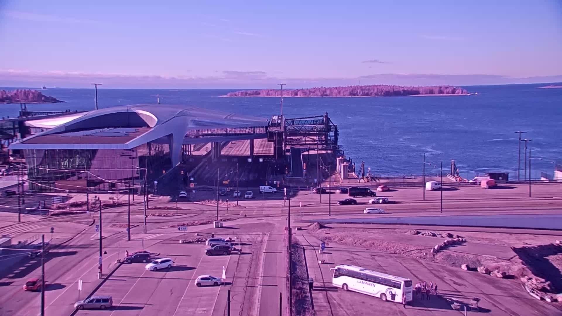 A modern ferry terminal with a curved roof sits by a body of water under a clear, sunny sky, with cars parked in lots and a bus parked nearby.