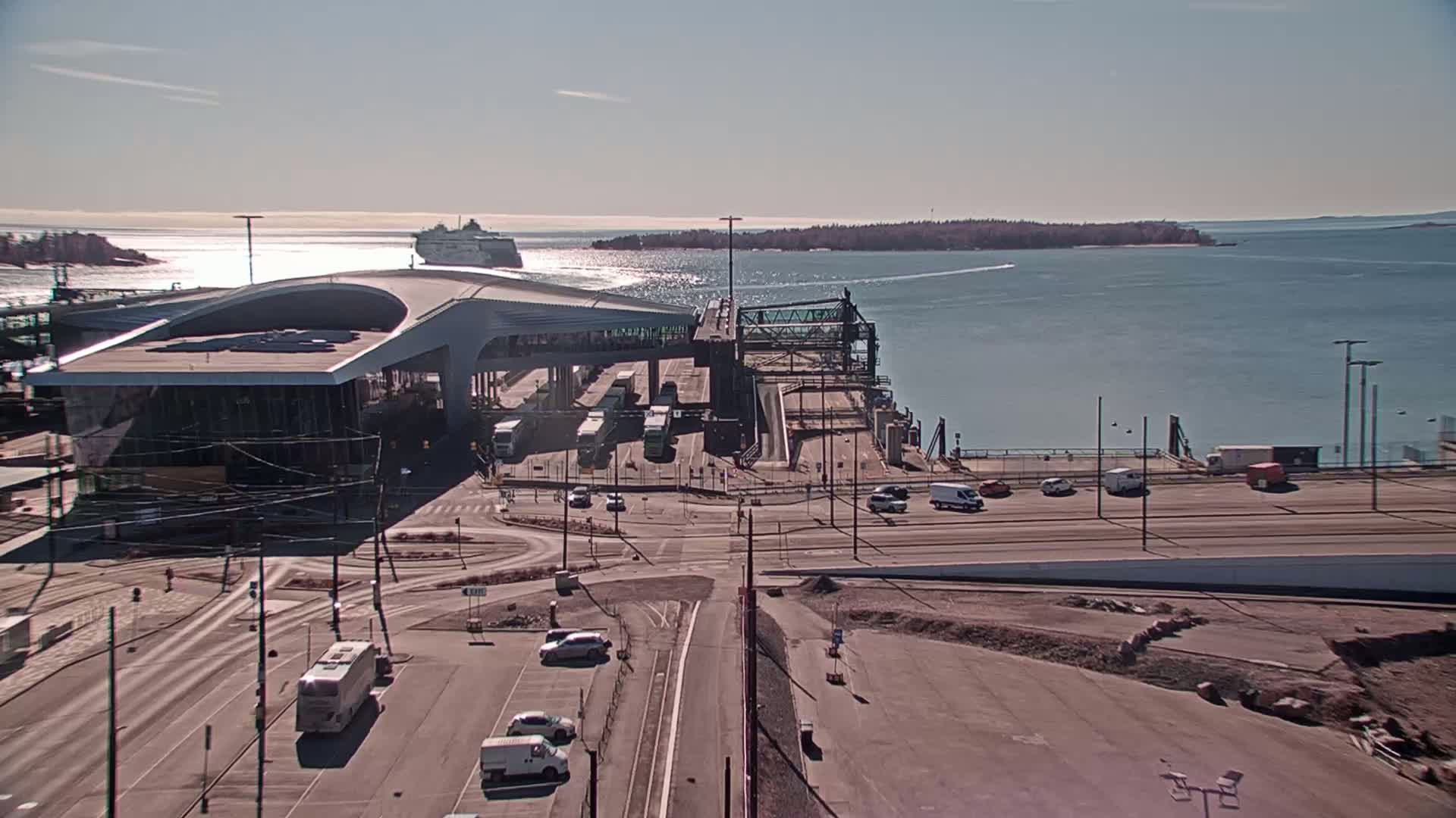 A ferry terminal with a large modern building, several vehicles, and a cruise ship in the distance on a sunny day.