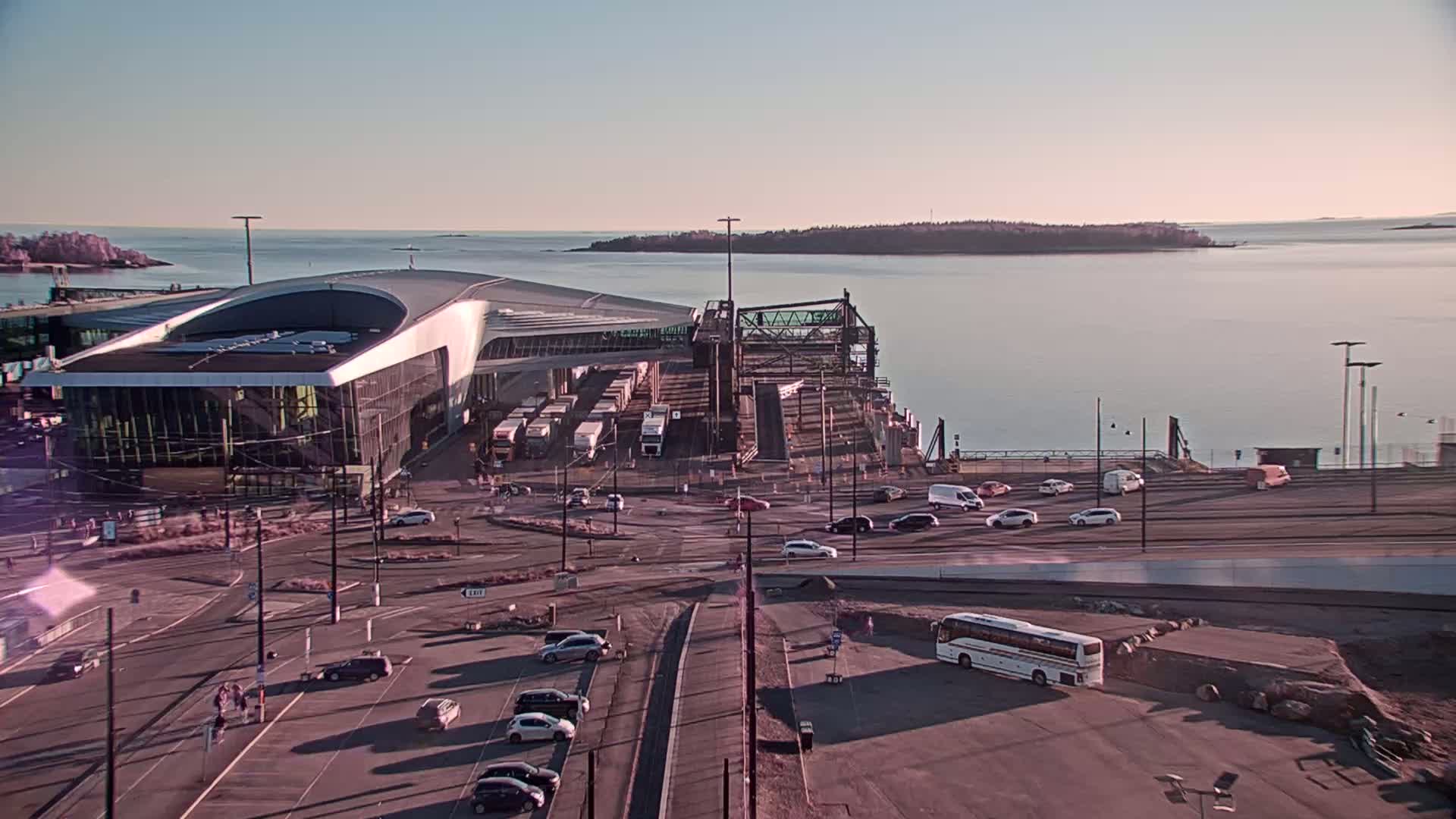 A ferry terminal with a modern building and numerous vehicles is situated by a calm body of water under a clear, pale sky.
