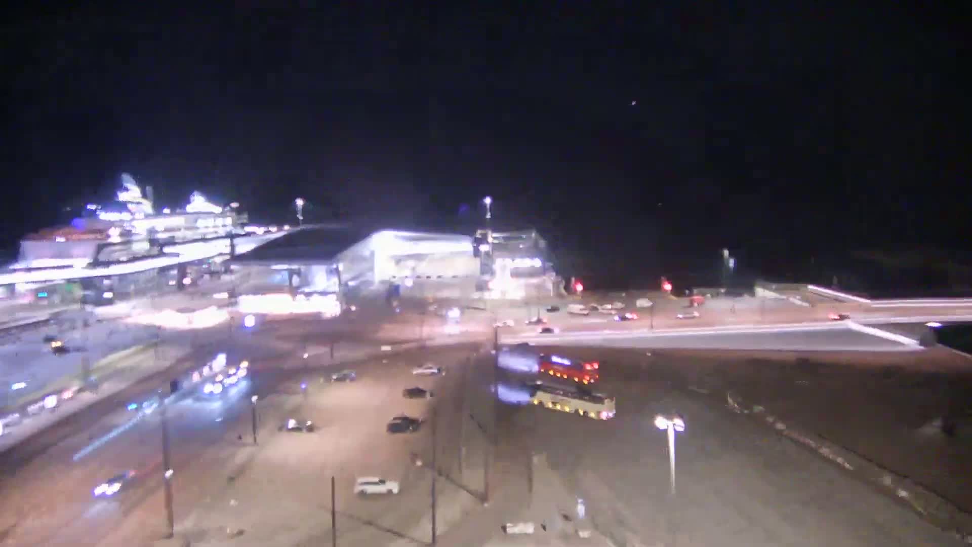 A nighttime, high-angle view shows a large cruise ship docked near a busy intersection with cars and a bus, all under a clear, dark sky.
