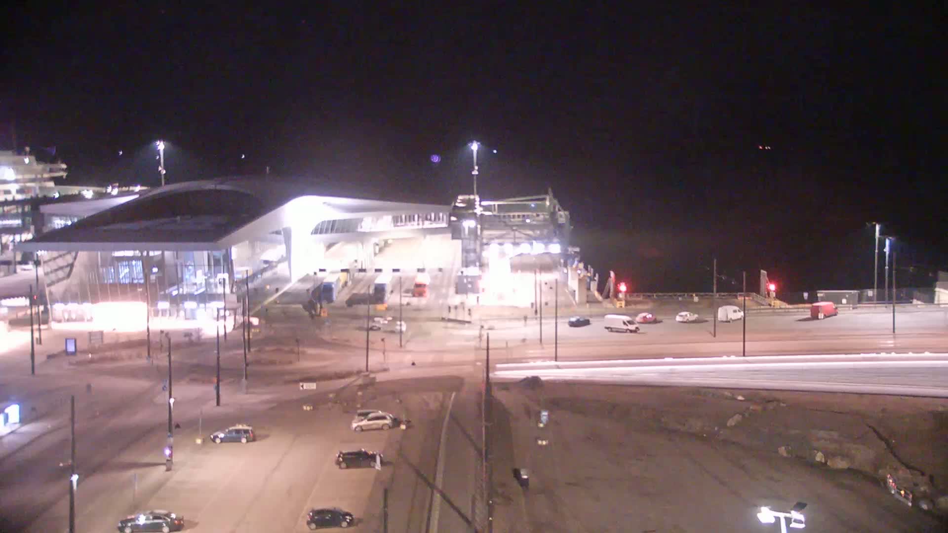 A ferry terminal at night with a few vehicles in the parking lot and some cars driving on the roads surrounding it, under a clear, dark sky.