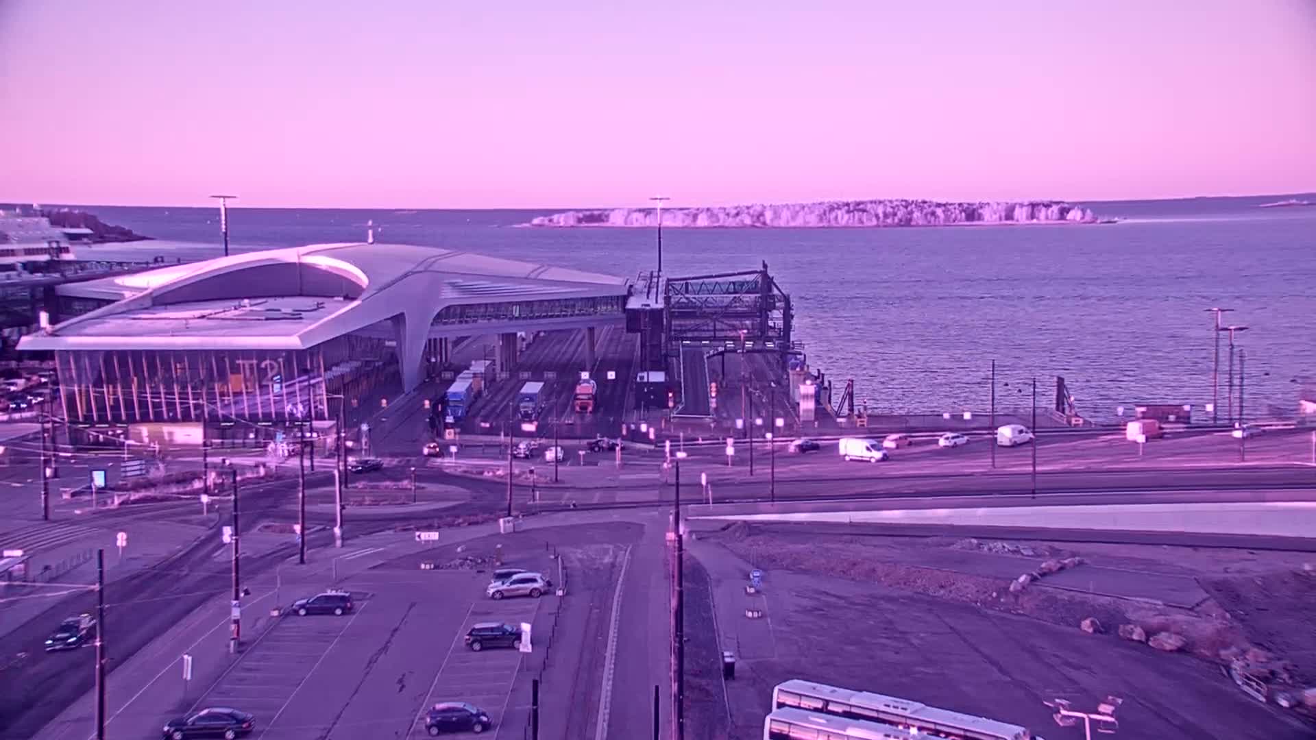 A ferry terminal with a modern building and several vehicles is situated by a body of water under a pale purple twilight sky, with an island visible in the distance.