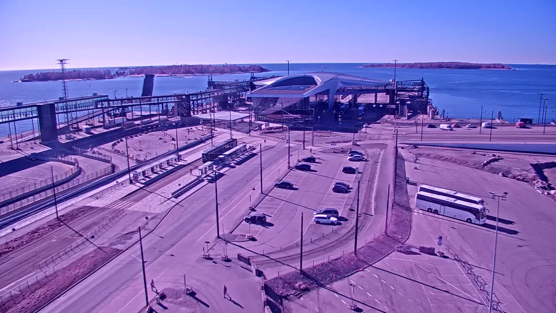 An aerial view shows a ferry terminal with a modern building, parking lots, a tram stop, and a bus parked nearby on a sunny day.