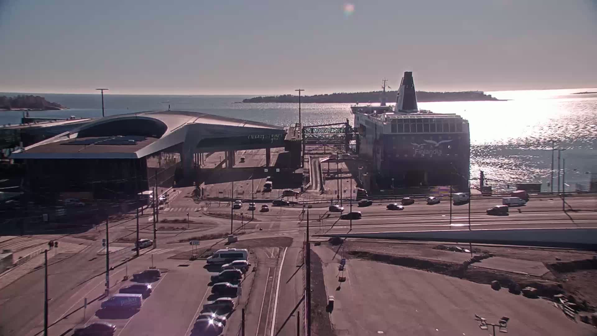 A ferry is docked at a terminal, with cars parked in lots nearby, under a sunny sky.