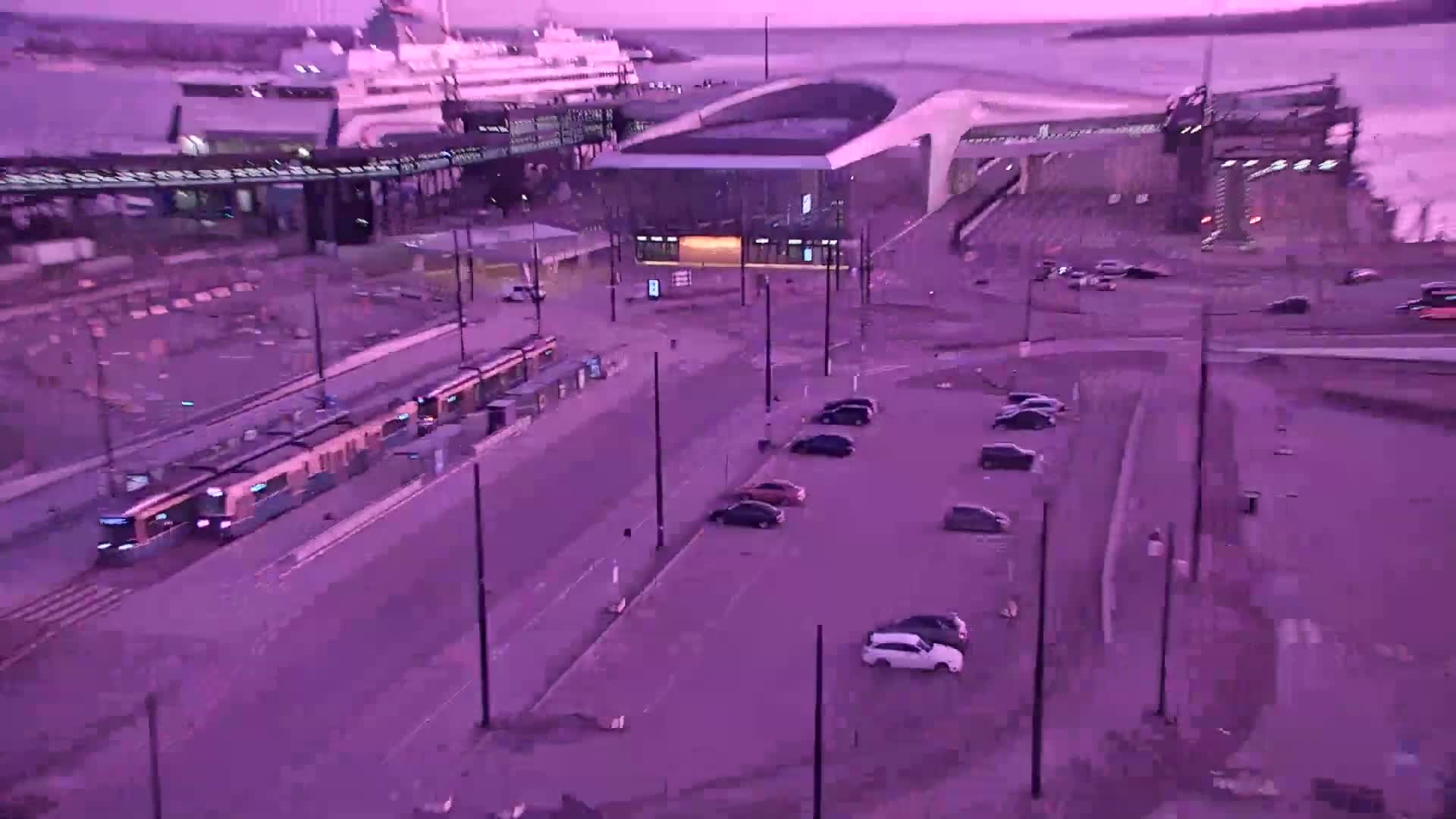 A high-angle view shows two trams at a station near a ferry terminal and parking lot under a purple twilight sky.