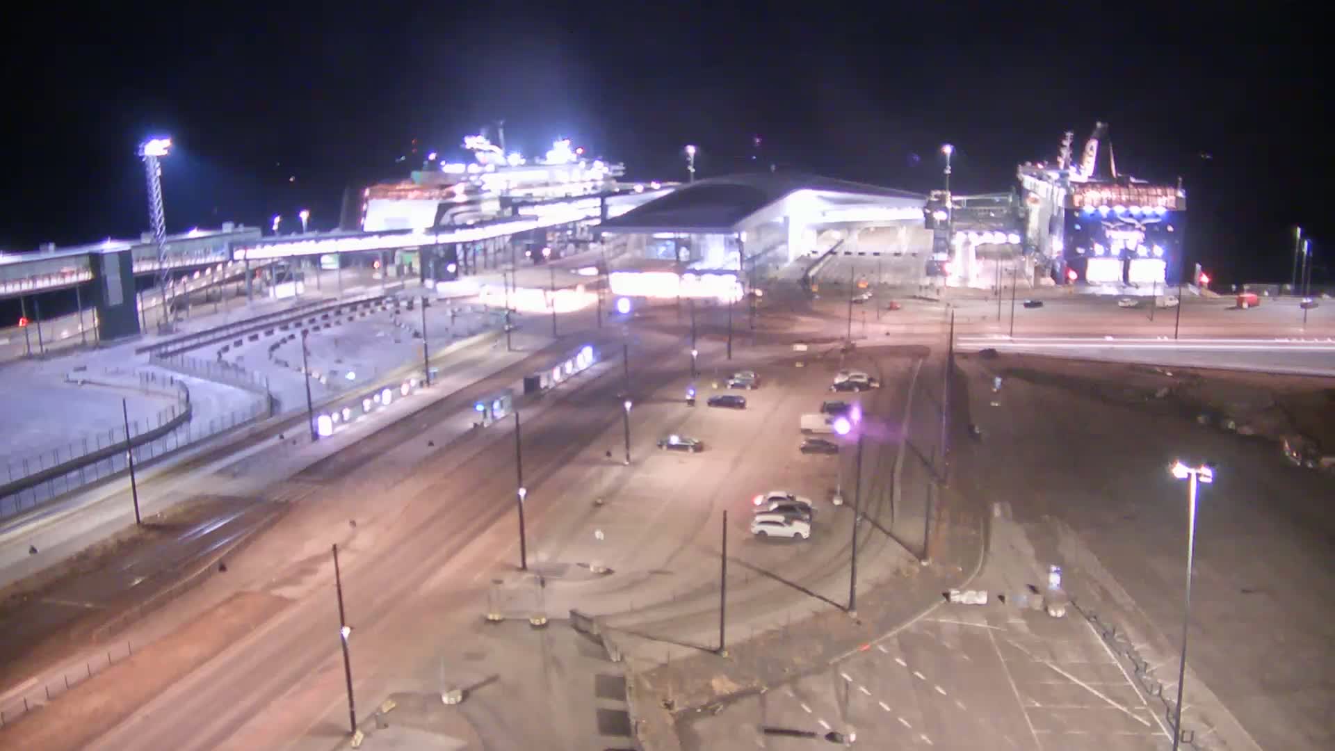 A nighttime aerial view shows a ferry terminal with two large ferries docked, a modern terminal building, and a sparsely populated parking lot.