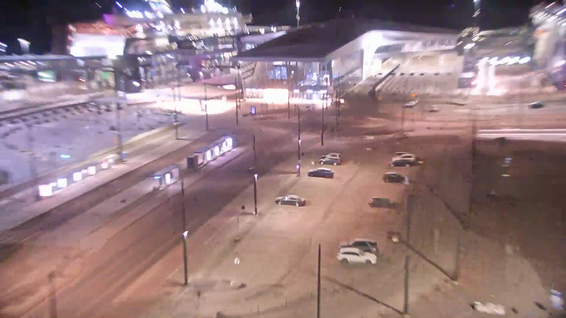 A snow-covered city street at night, with cars parked and a train track visible, next to a large building.