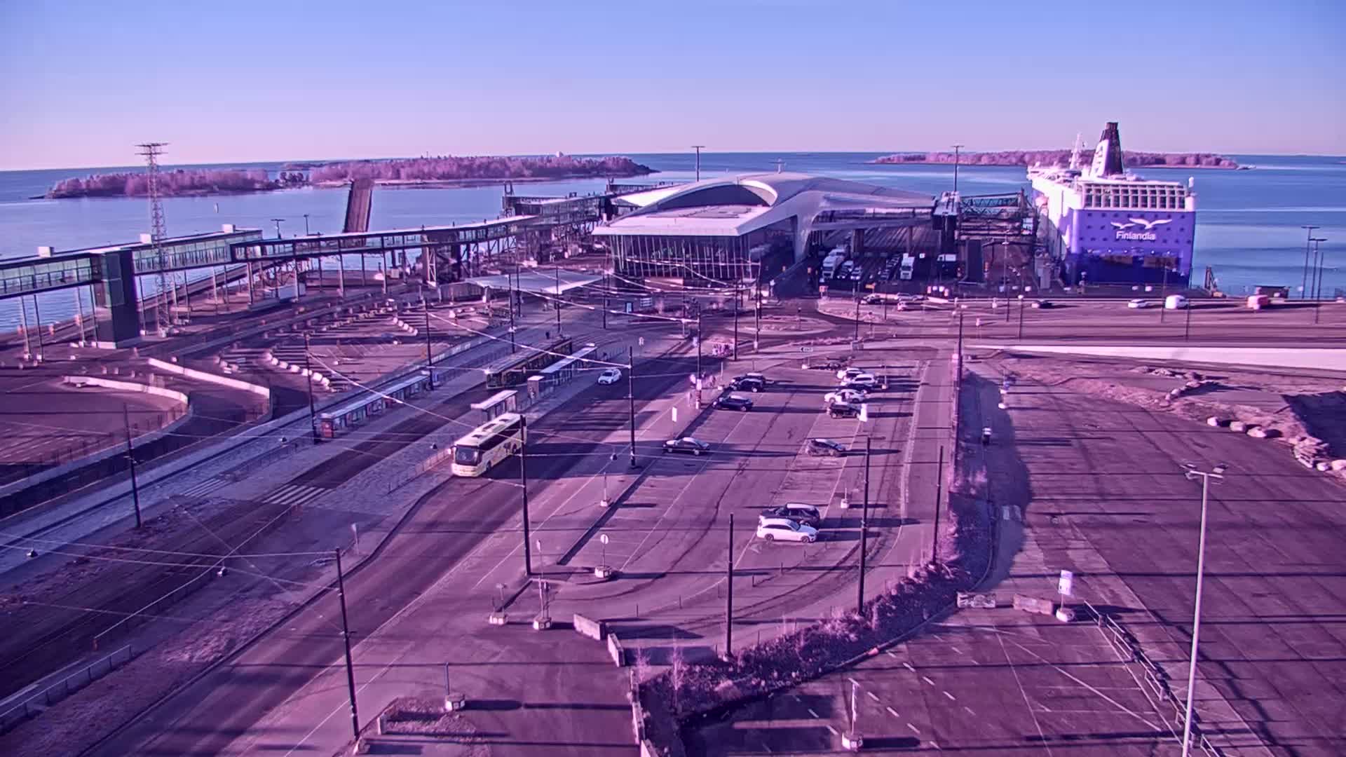A ferry is docked at a terminal on a clear, sunny day, with trams and cars visible in a nearby parking lot.