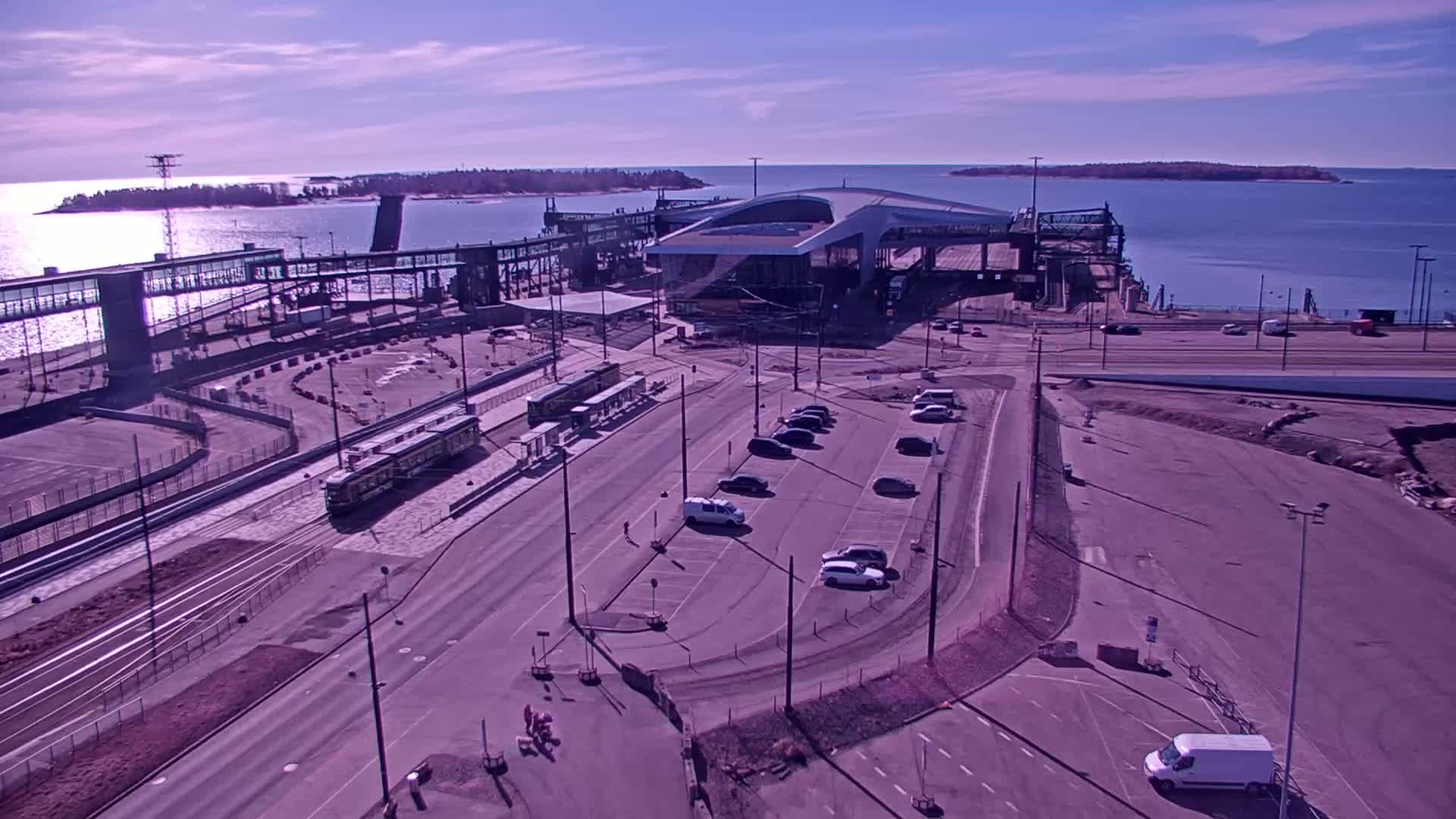 An aerial view shows a ferry terminal with trams and parked cars on a sunny day, with calm water and islands visible in the background.