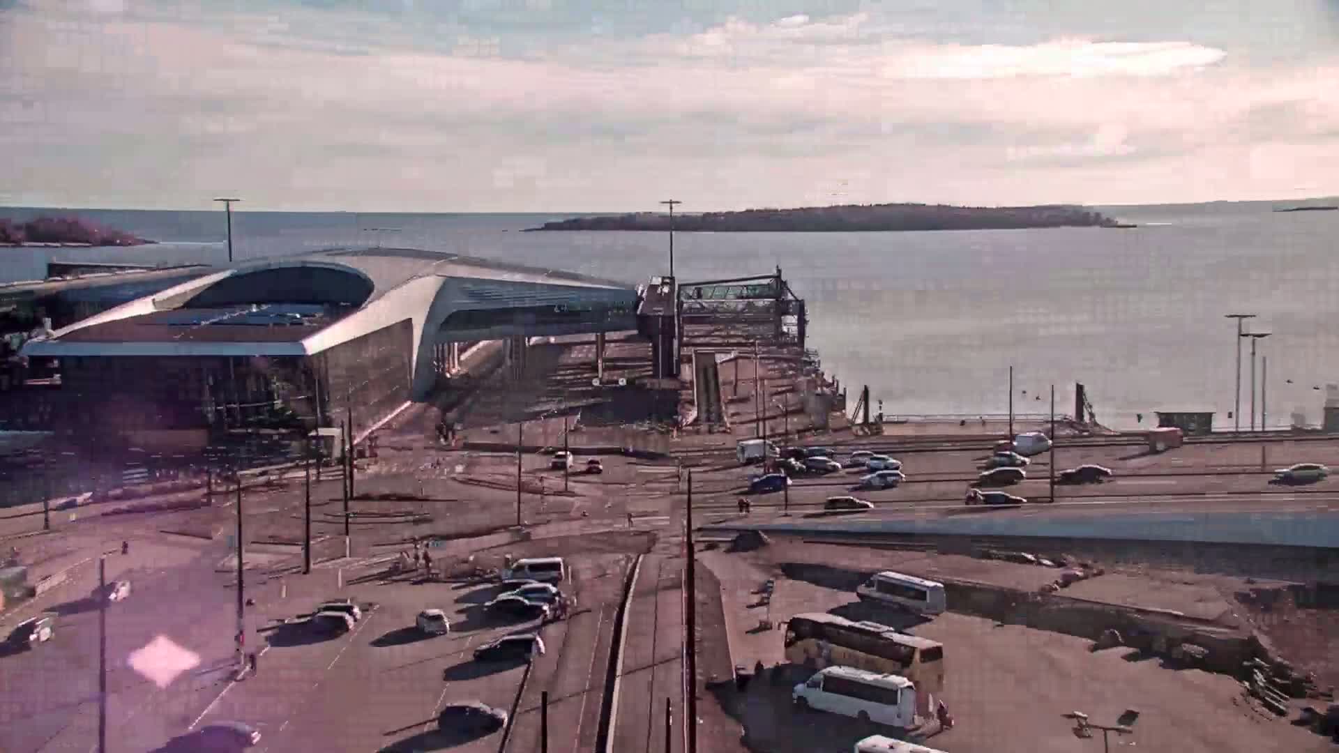 A modern ferry terminal with cars and buses in a parking lot next to it overlooks a calm body of water on a partly sunny day.