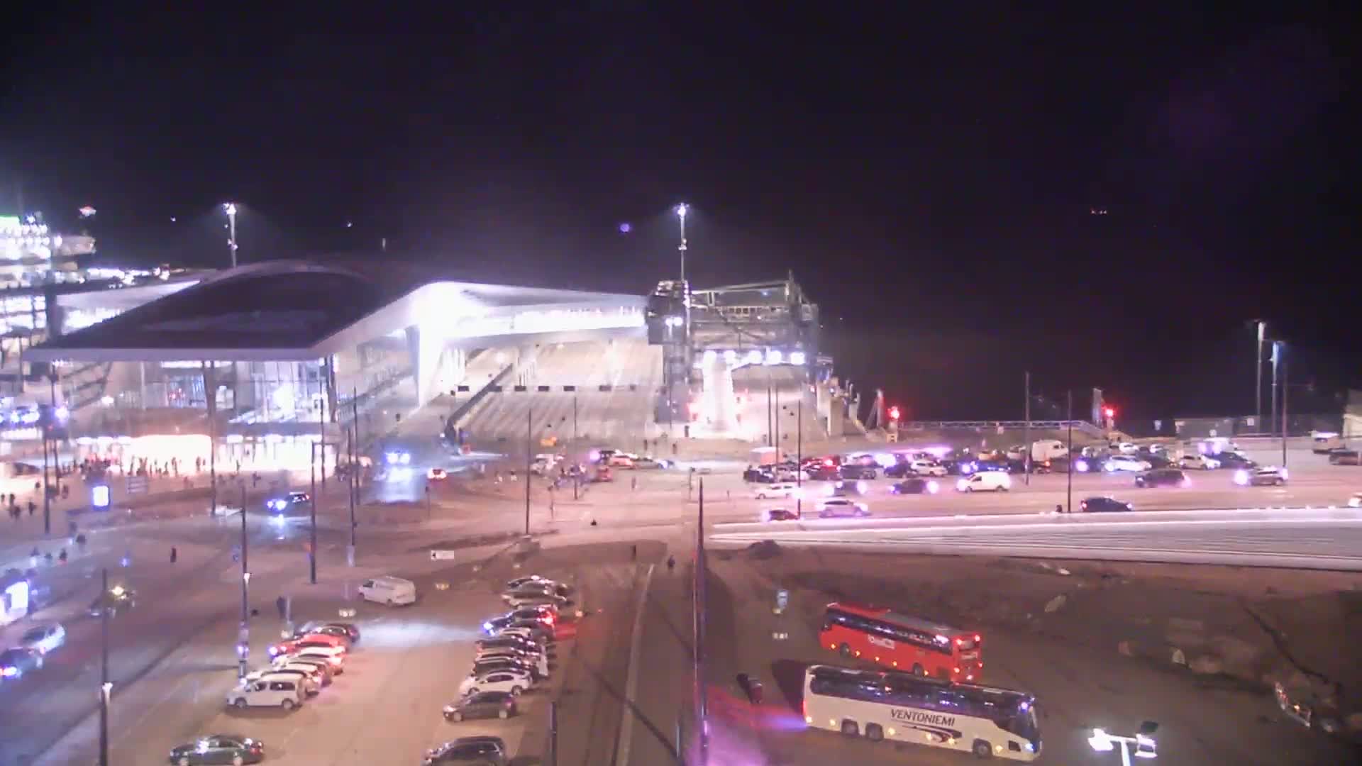 At night, a brightly lit stadium and surrounding area are filled with numerous cars and buses, with some emergency vehicles present.