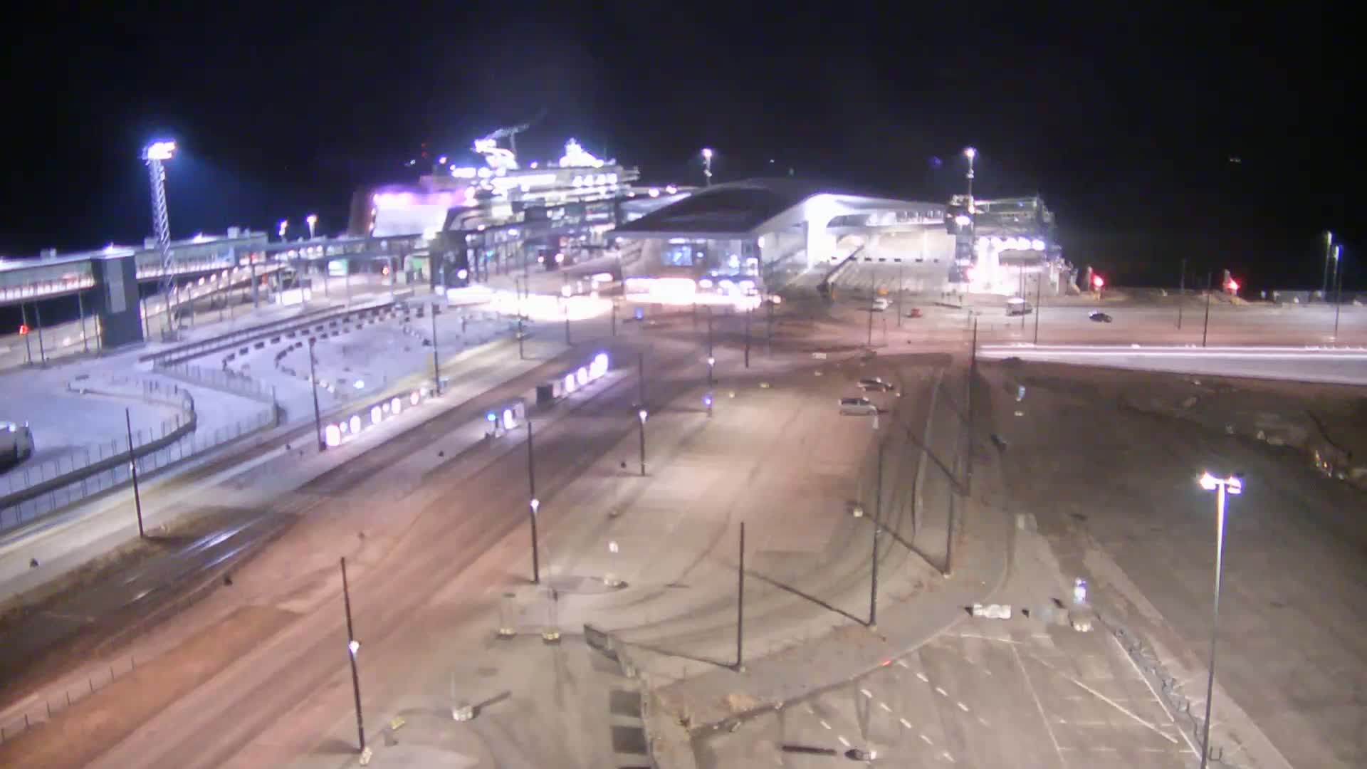 A nighttime aerial view of a port area shows a large cruise ship docked near a terminal building and surrounding roadways, with some light snow or ice on the ground.
