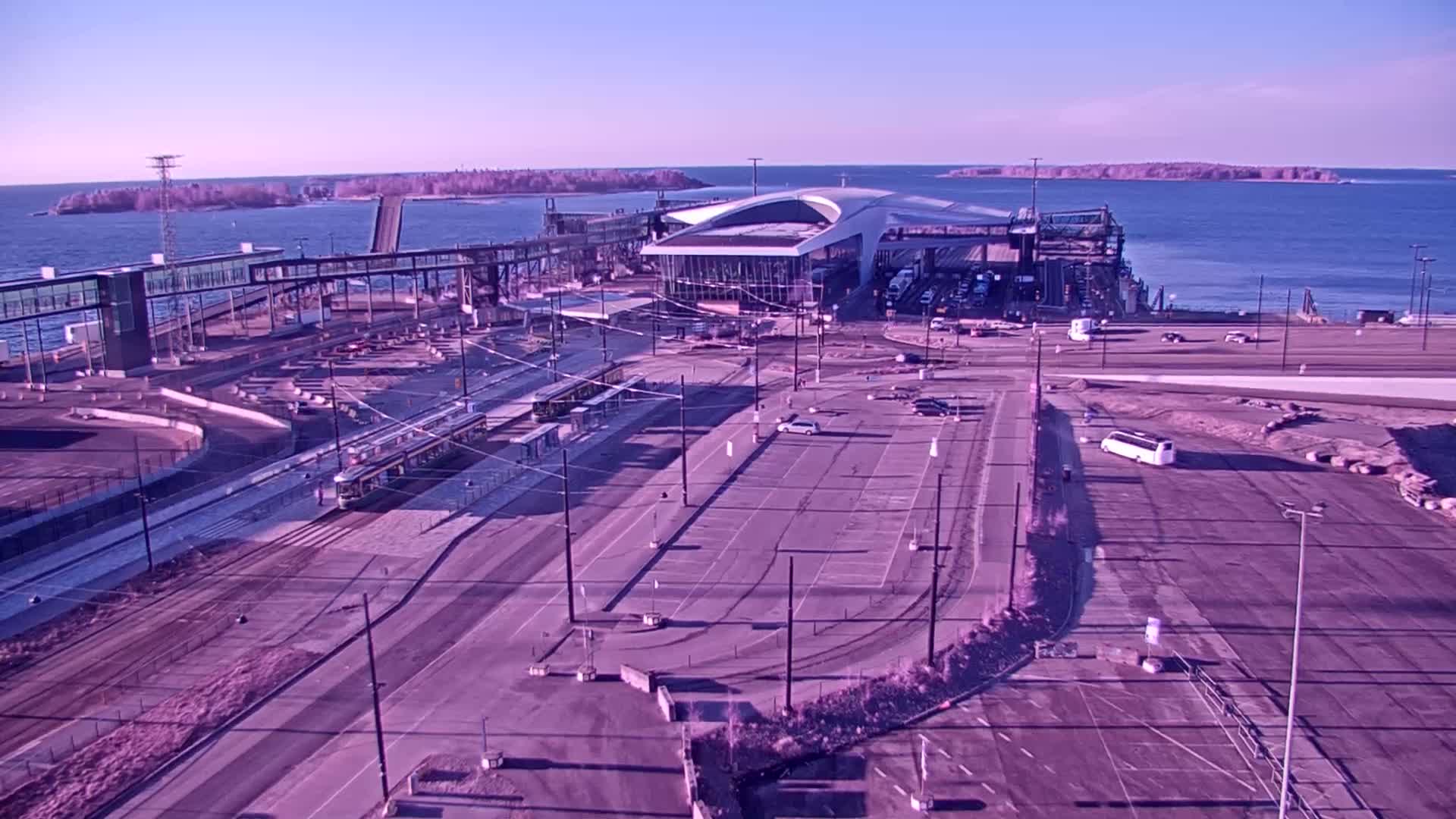 An aerial view of a ferry terminal on a clear, bright day shows a large terminal building, several vehicles, and trams on tracks, with calm water and islands visible in the background.
