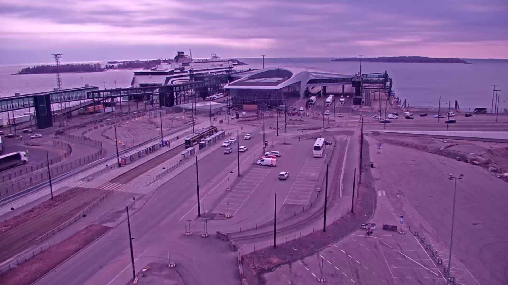 An aerial view of a ferry terminal with a large ferry docked, several vehicles in a parking lot, and a tram at a station, under a dusky purple sky.